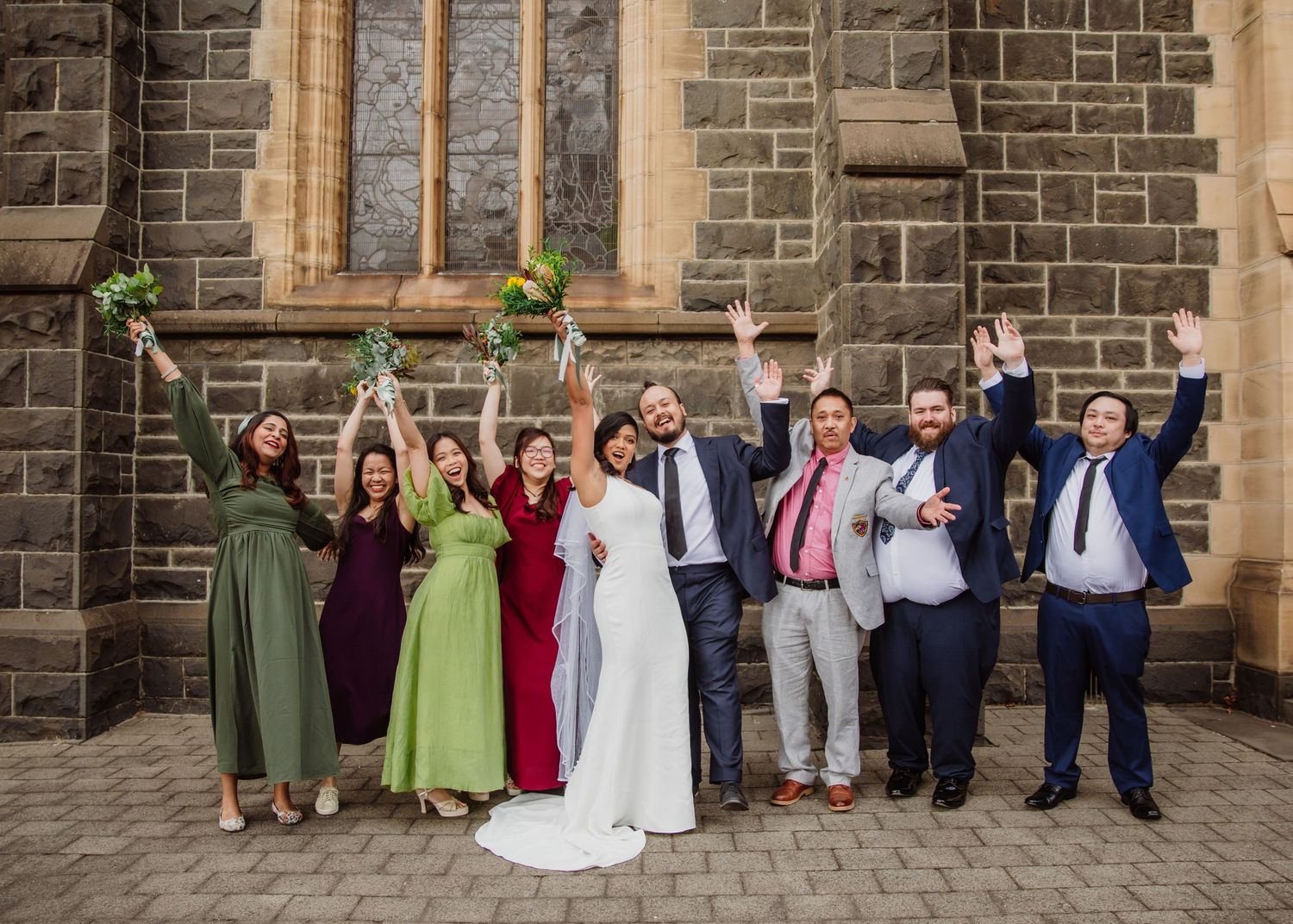 Joyful wedding party celebrates outside a stone church with bridesmaids in colorful dresses and groomsmen cheering.