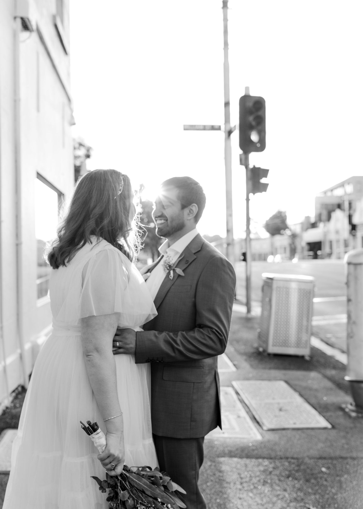 Black and white photo of smiling bride and groom embracing on urban street corner at sunset.
