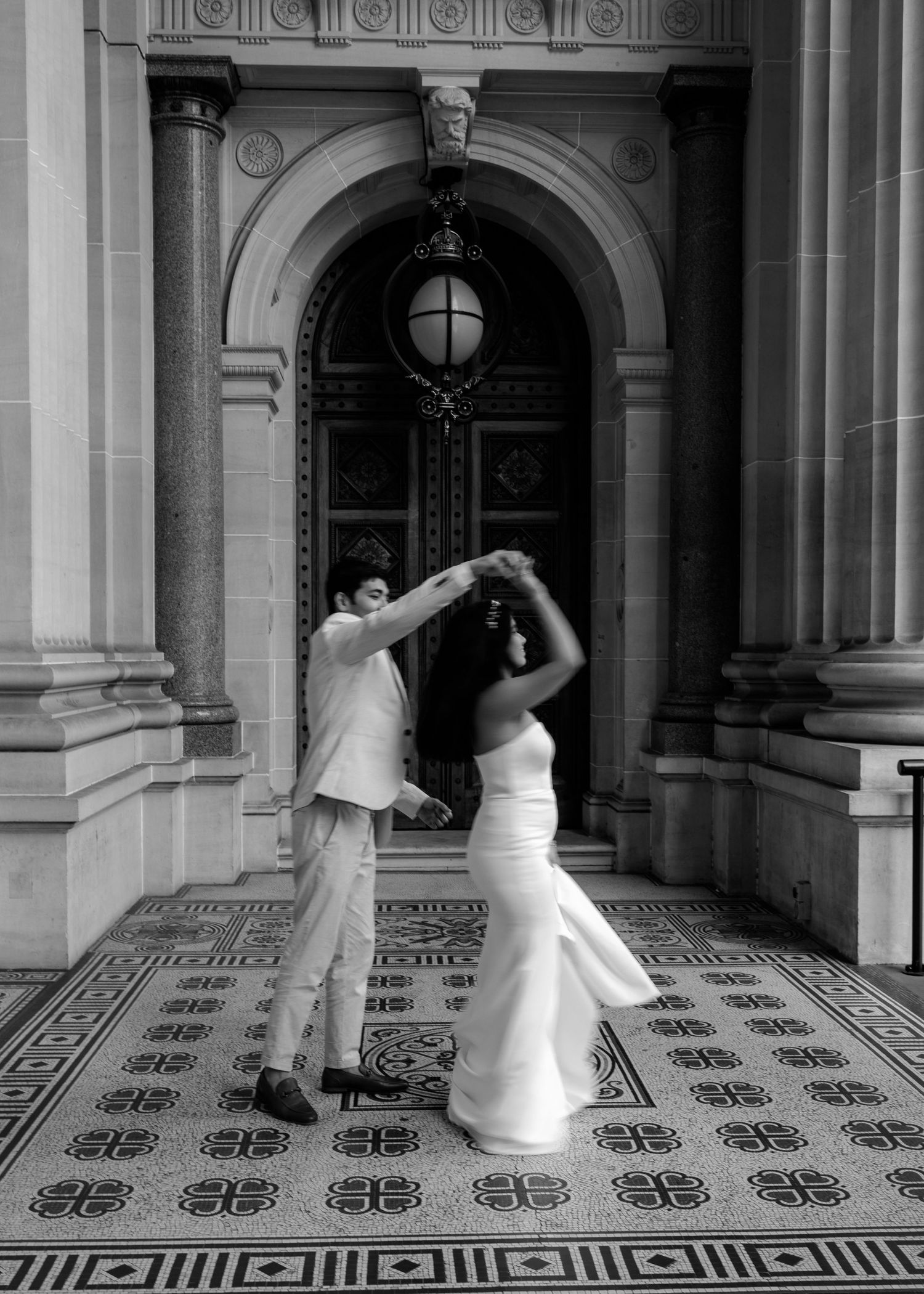 Black and white photo of bride and groom dancing elegantly in front of ornate arched doorway on decorative tiled floor.