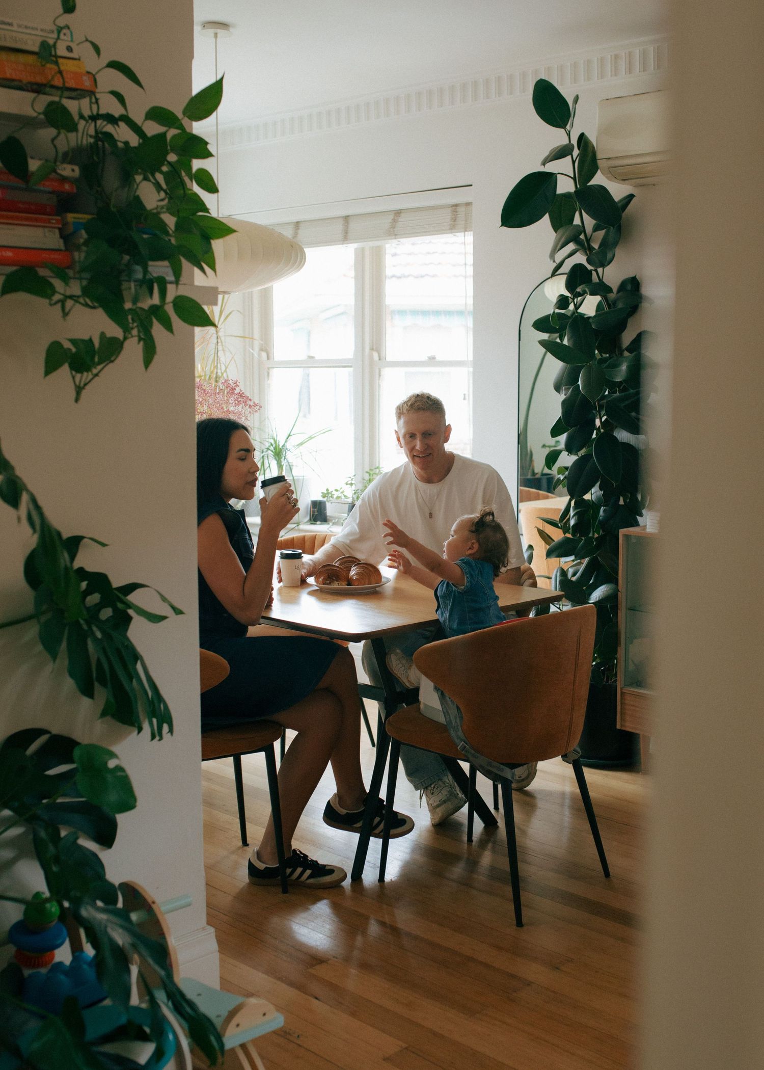 Family with toddler sharing breakfast at dining table in plant-filled bright home interior.