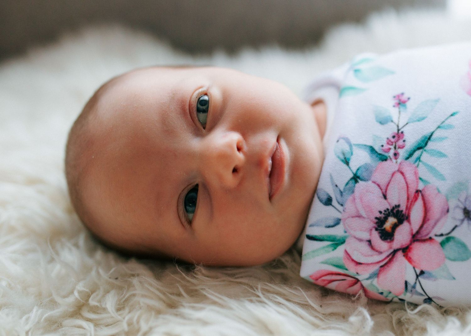 Newborn baby with blue eyes lying on a white fur rug in a floral swaddle.
