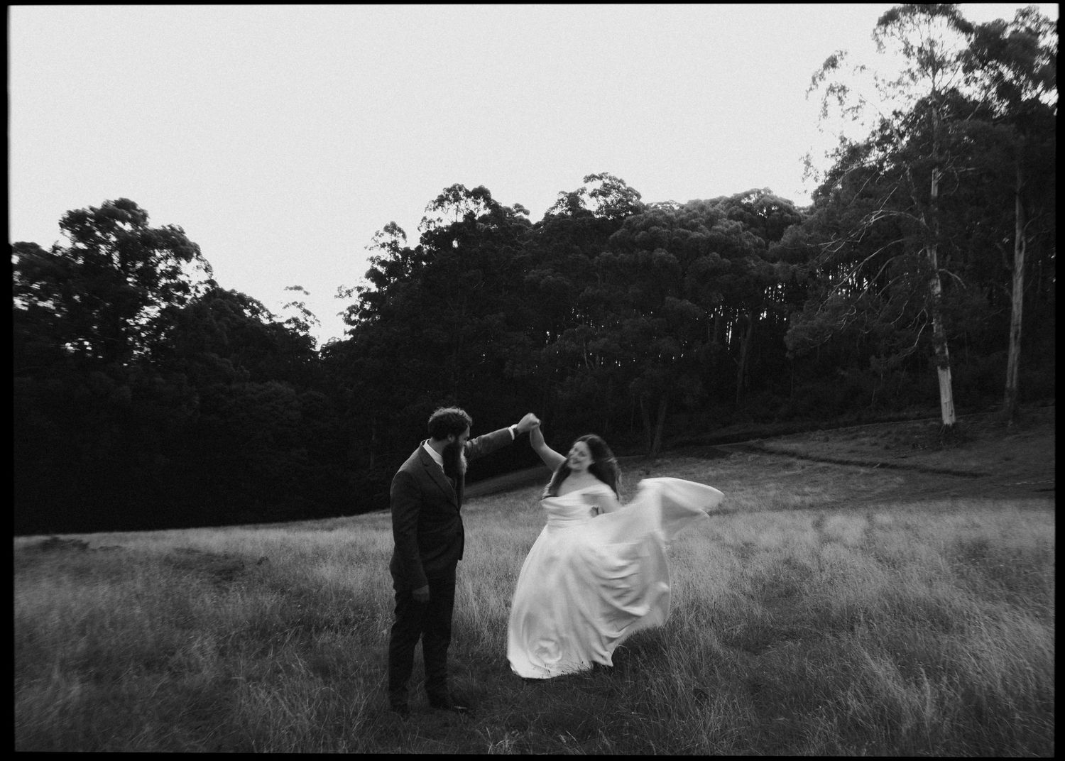 Black and white photo of groom twirling bride in a flowing dress in a field at dusk.