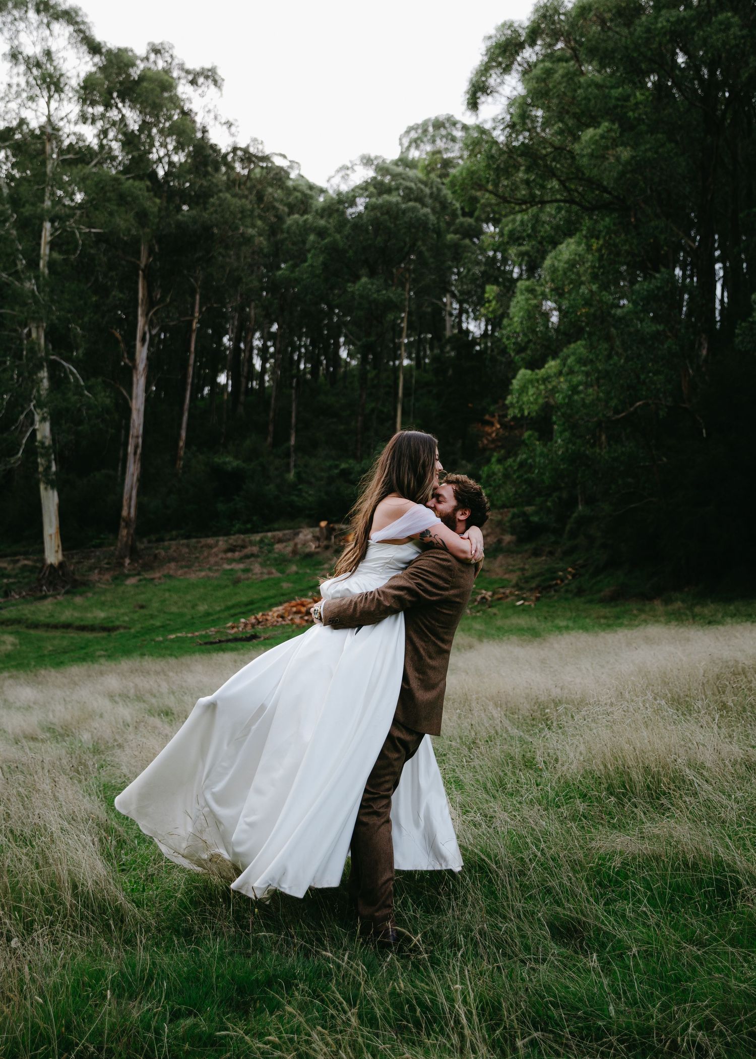 Groom lifting bride in a flowing white dress in a scenic meadow surrounded by tall eucalyptus trees.
