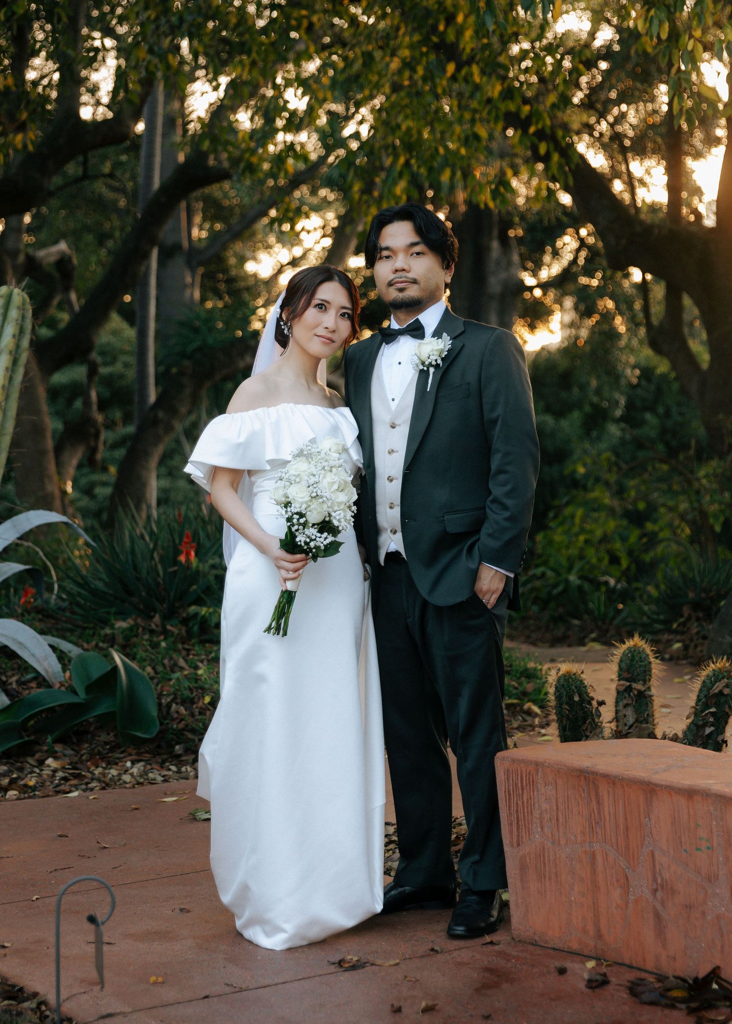 Bride and groom portrait in garden, she wears off-shoulder gown holding white bouquet.
