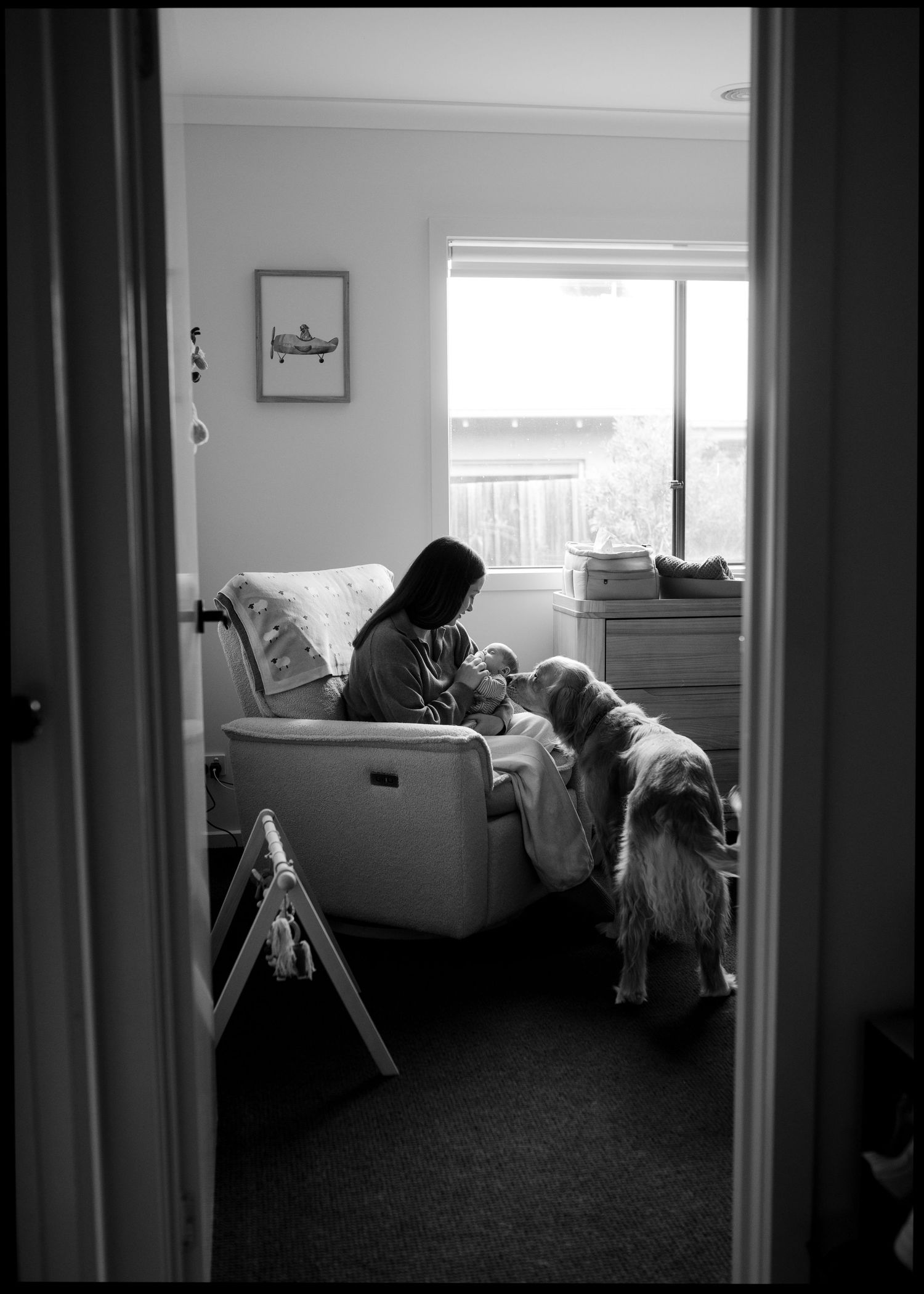 Black and white photo of a mother holding her newborn in a nursery chair while a golden retriever looks on.