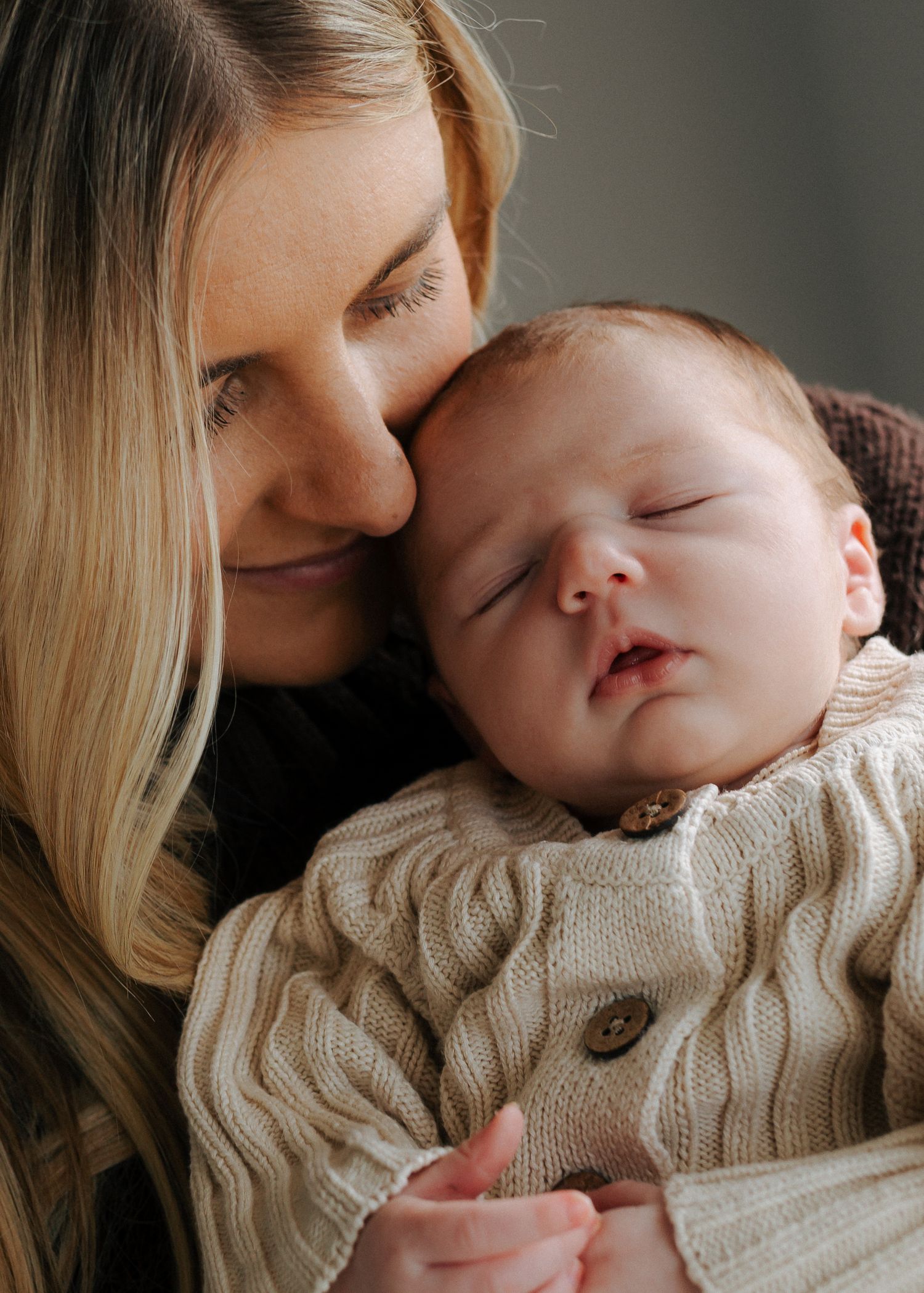 Close-up of blonde mother cheek to cheek with sleeping newborn dressed in cream knit cardigan.