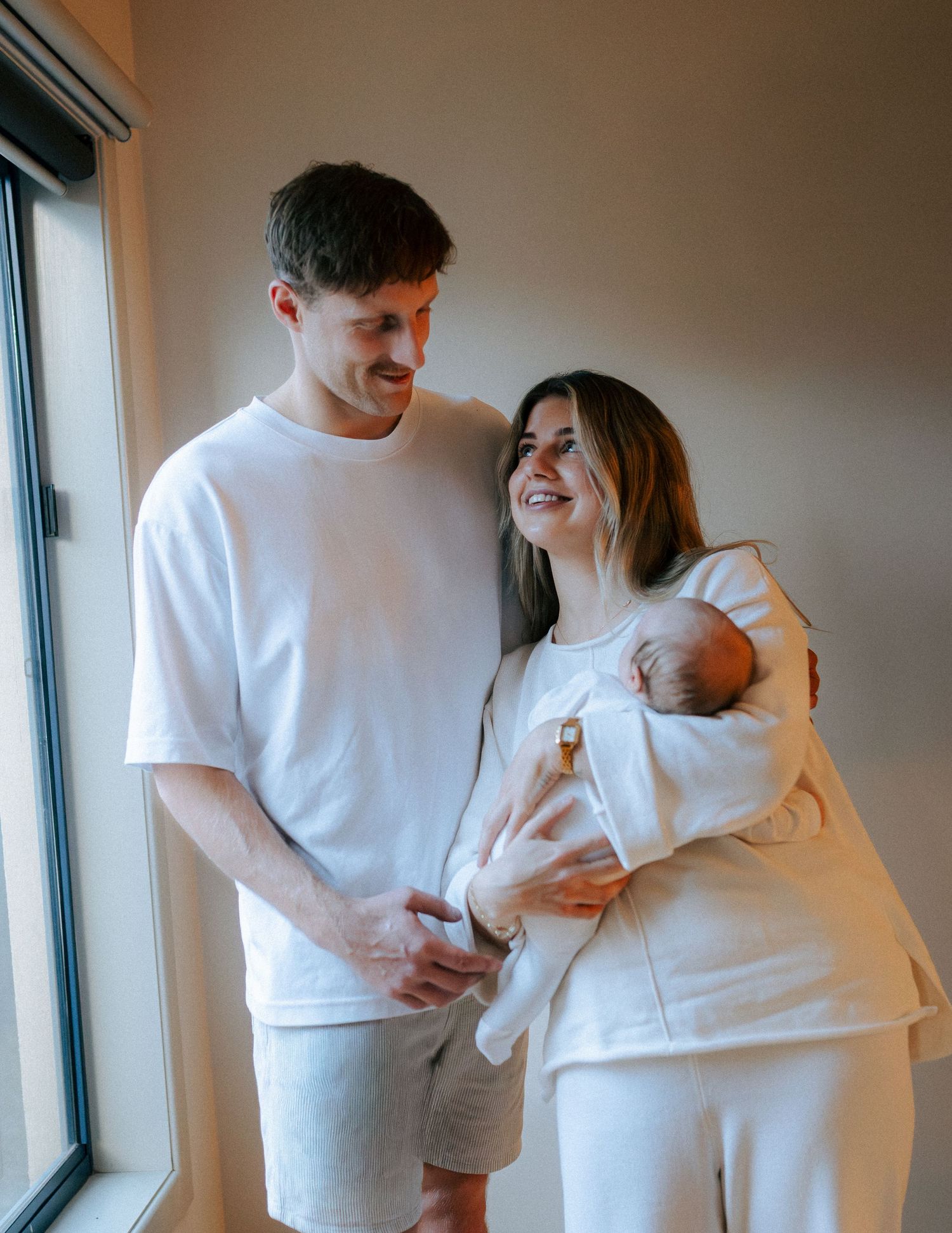 Smiling new parents in white outfits holding newborn baby together near a bright window.