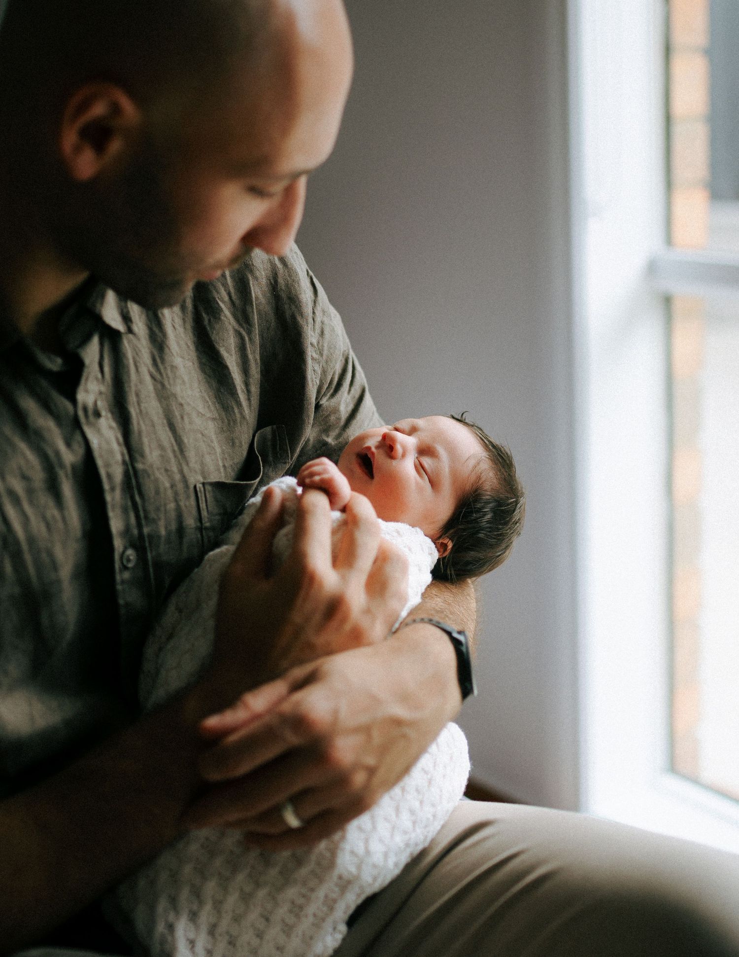 Bald father holding wide-eyed newborn baby near bright window in tender intimate moment.