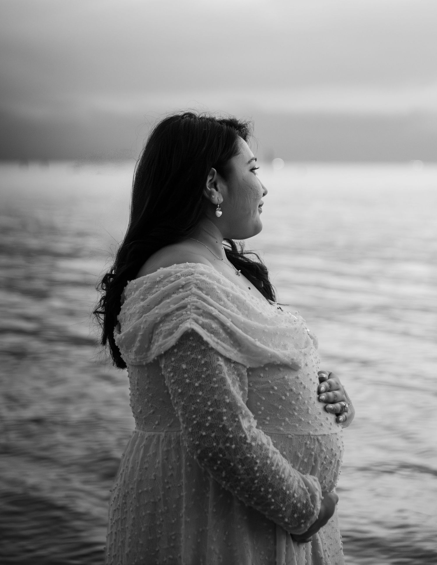 Black and white maternity photo of pregnant woman in white lace dress holding belly by the ocean.