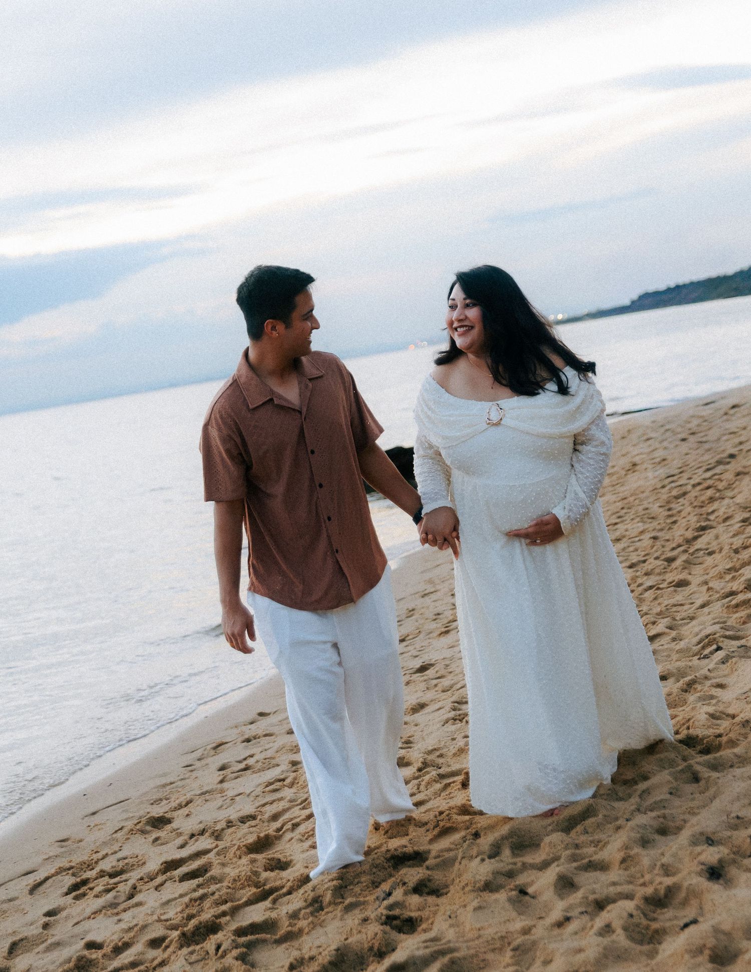 Smiling pregnant couple walking hand in hand along a sandy beach at sunset.