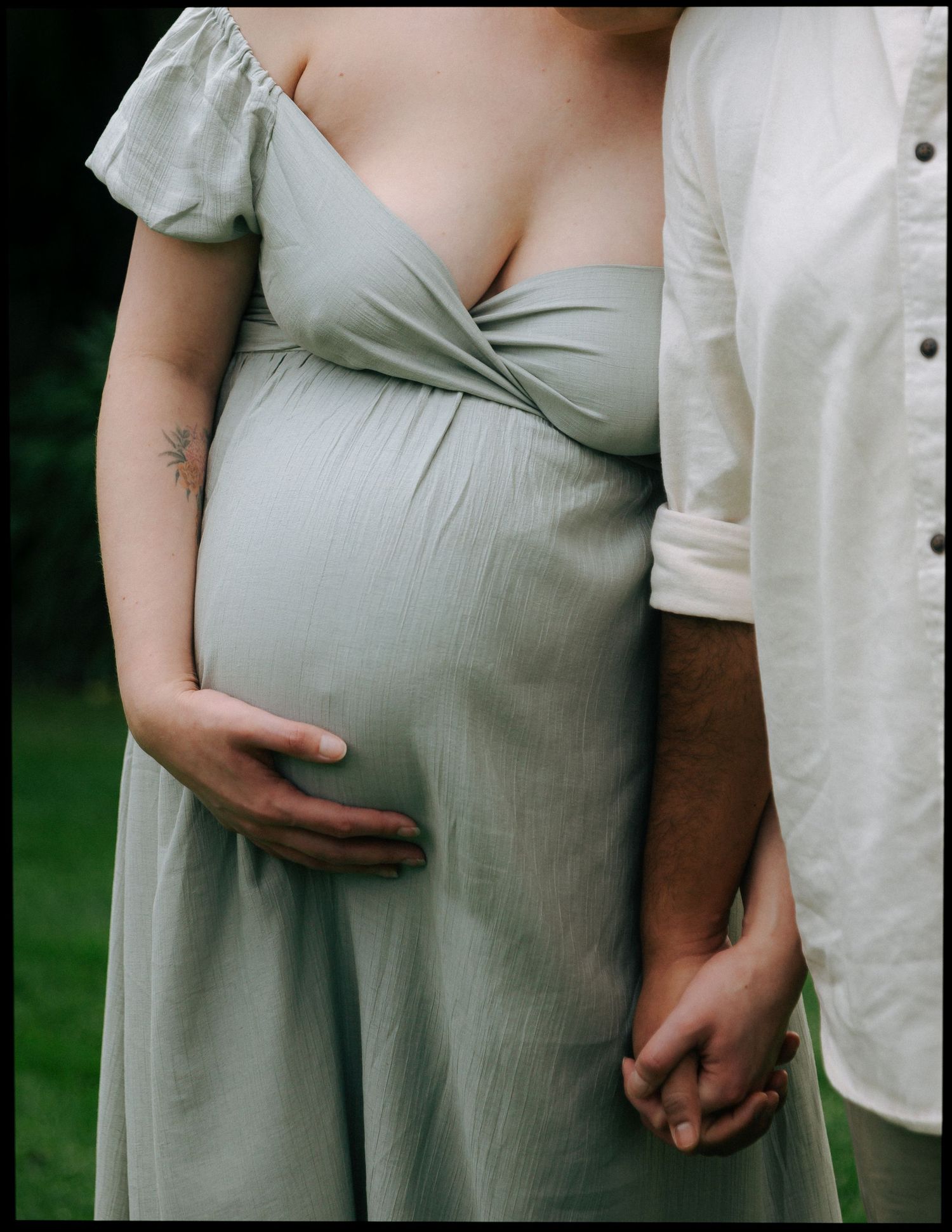 Close-up of pregnant woman in sage dress holding hands with partner, baby bump prominently displayed.