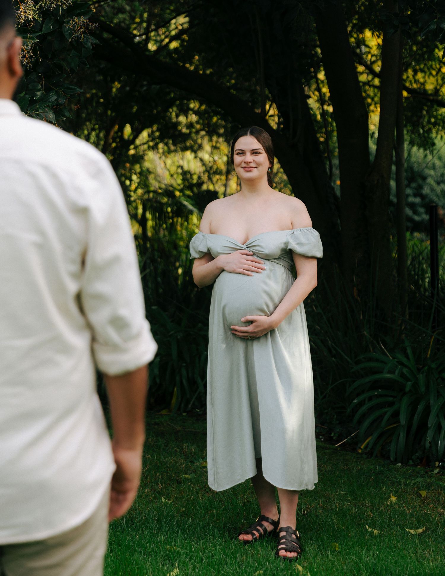 Pregnant woman in sage off-shoulder dress smiling and cradling belly while partner approaches in sunny garden.