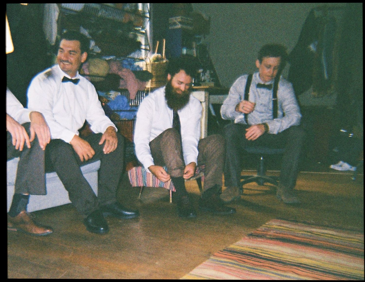 Three groomsmen getting ready together in a rustic room, adjusting bow ties and shoes.