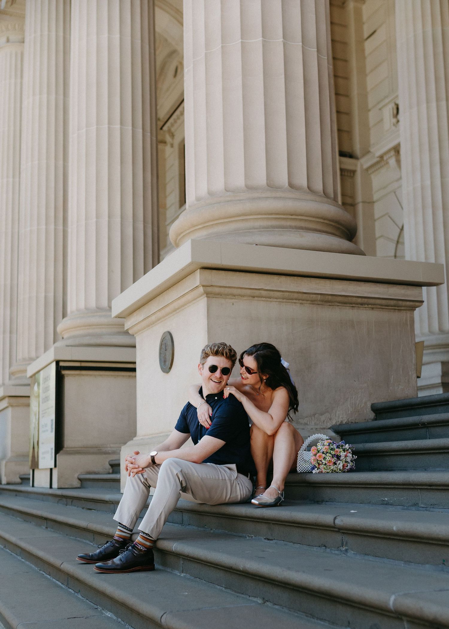Couple sitting on steps of a grand neoclassical building, sharing a tender moment beside large columns.