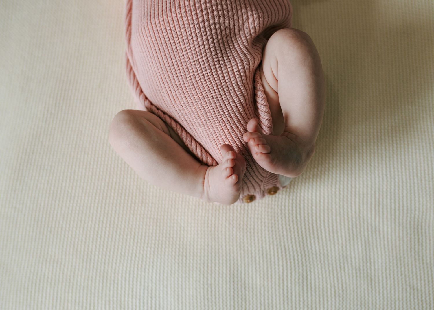Newborn baby in pink ribbed outfit lying on cream blanket with feet curled up.