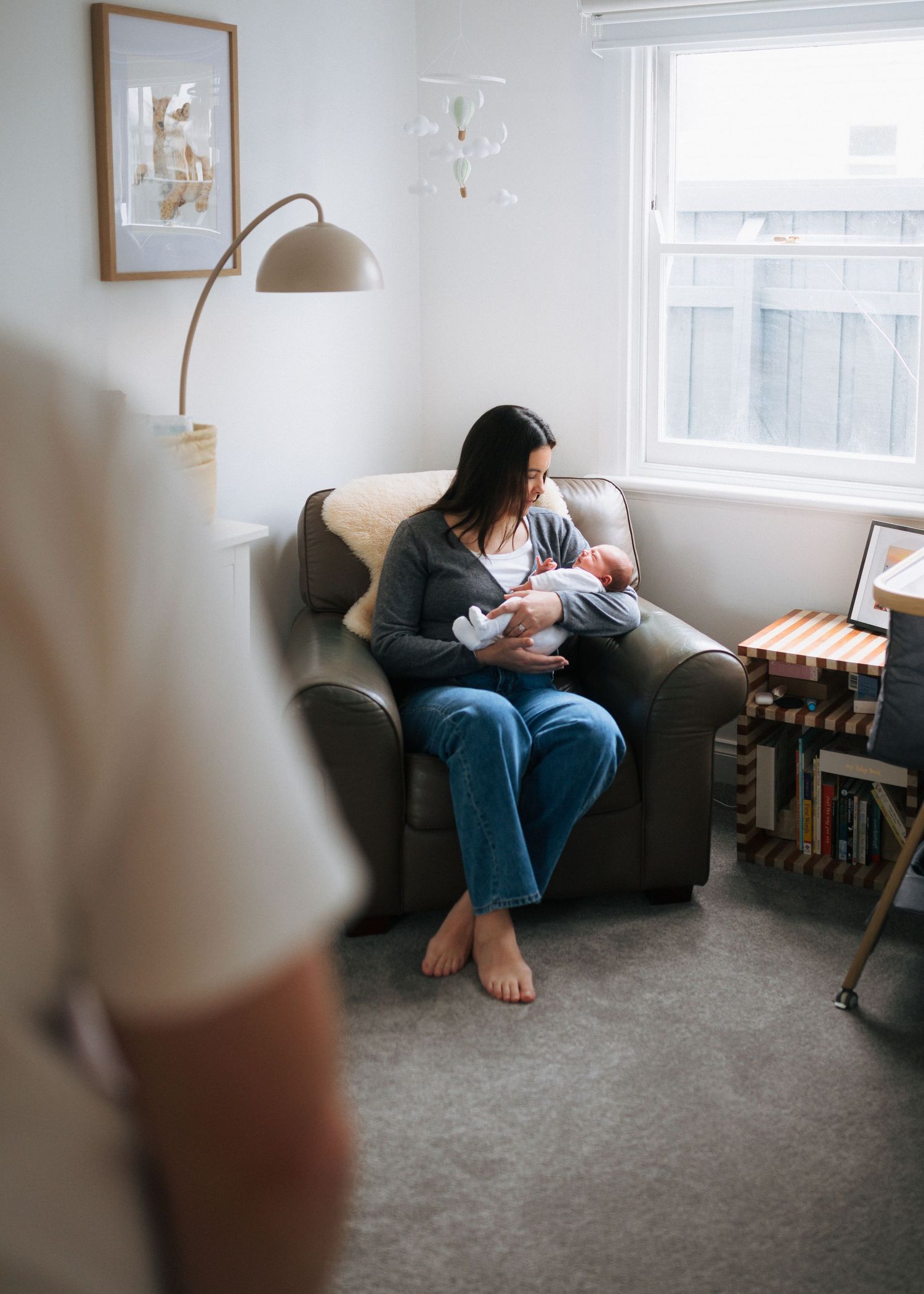 Barefoot mother in jeans sits in a leather armchair holding her newborn baby in a calm, softly lit nursery with a window and bookshelf.