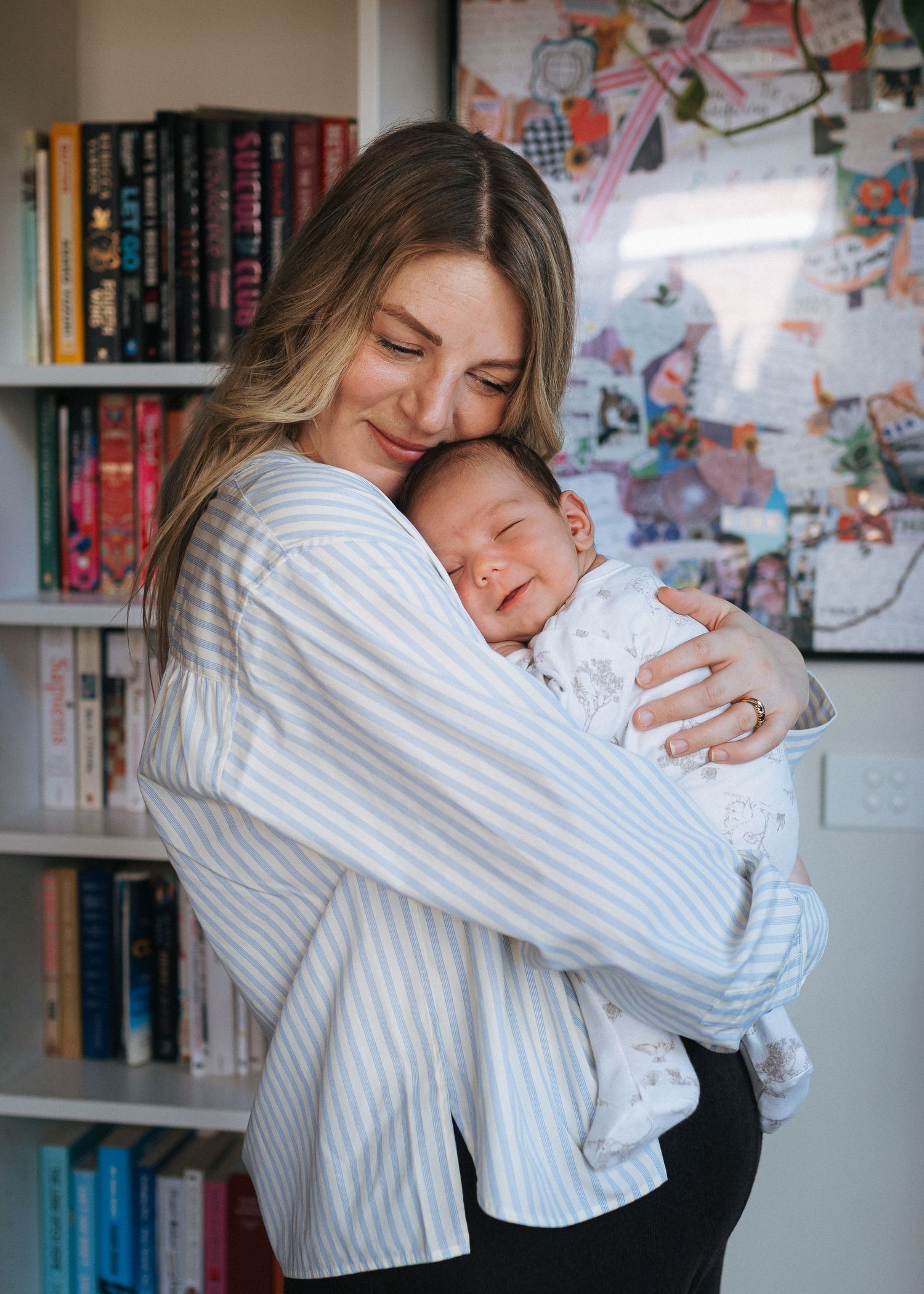 A smiling woman in a striped button-up shirt gently cradles a sleeping newborn baby in front of a bookshelf and a colorful collage board.