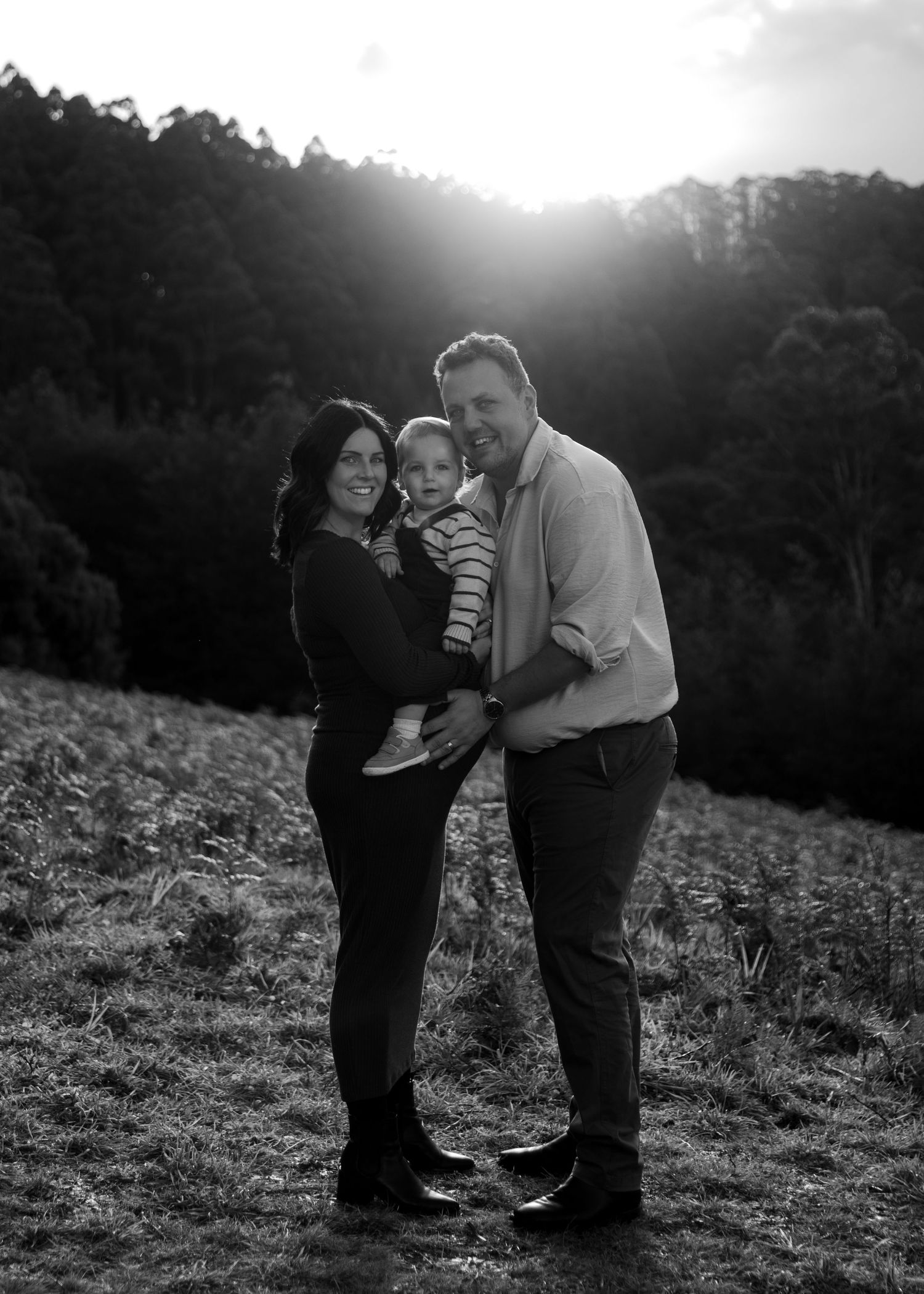 Black and white portrait of smiling family with toddler backlit by sunset on hillside.