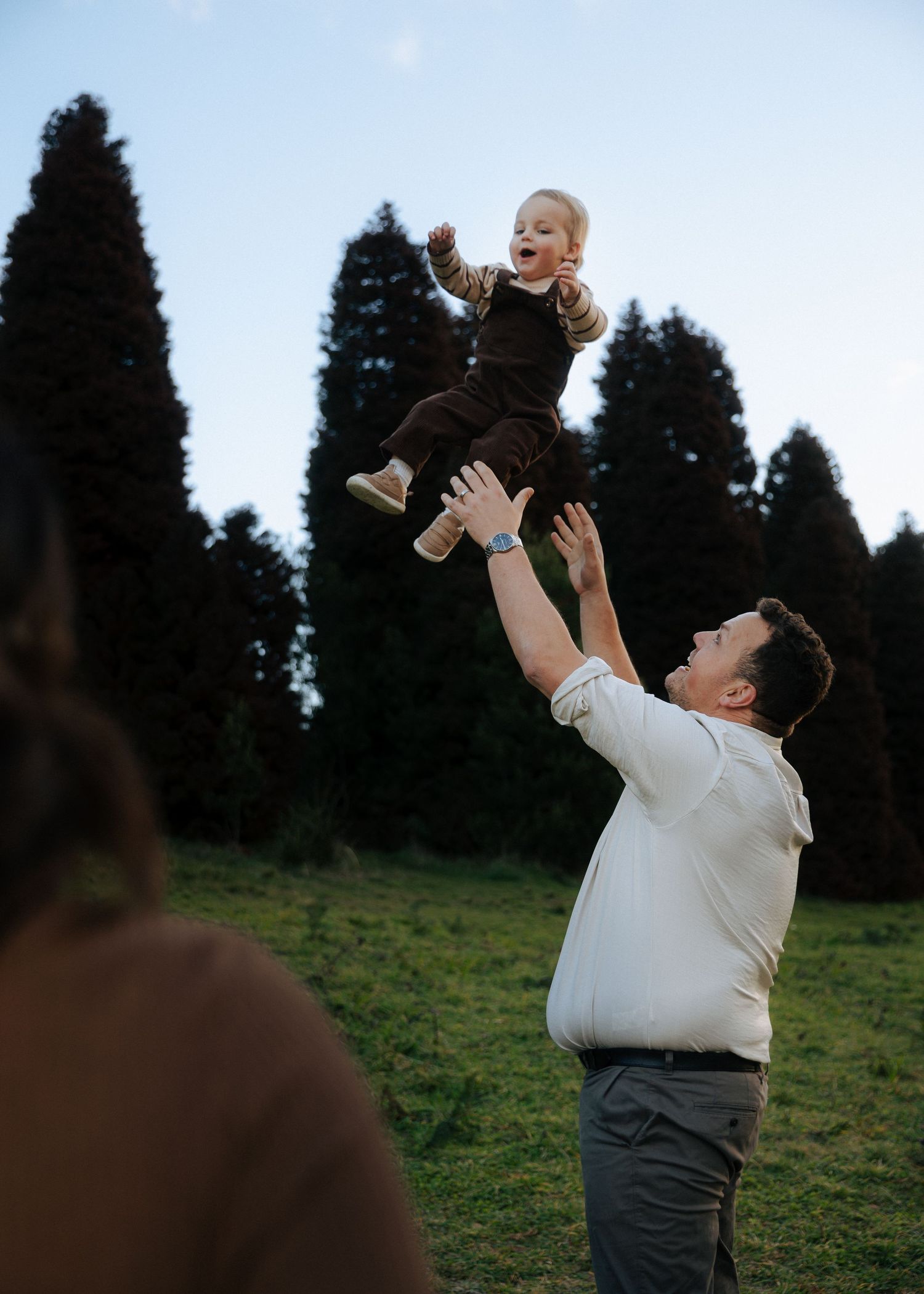 Man tossing laughing toddler in the air in front of tall trees during outdoor family photoshoot.