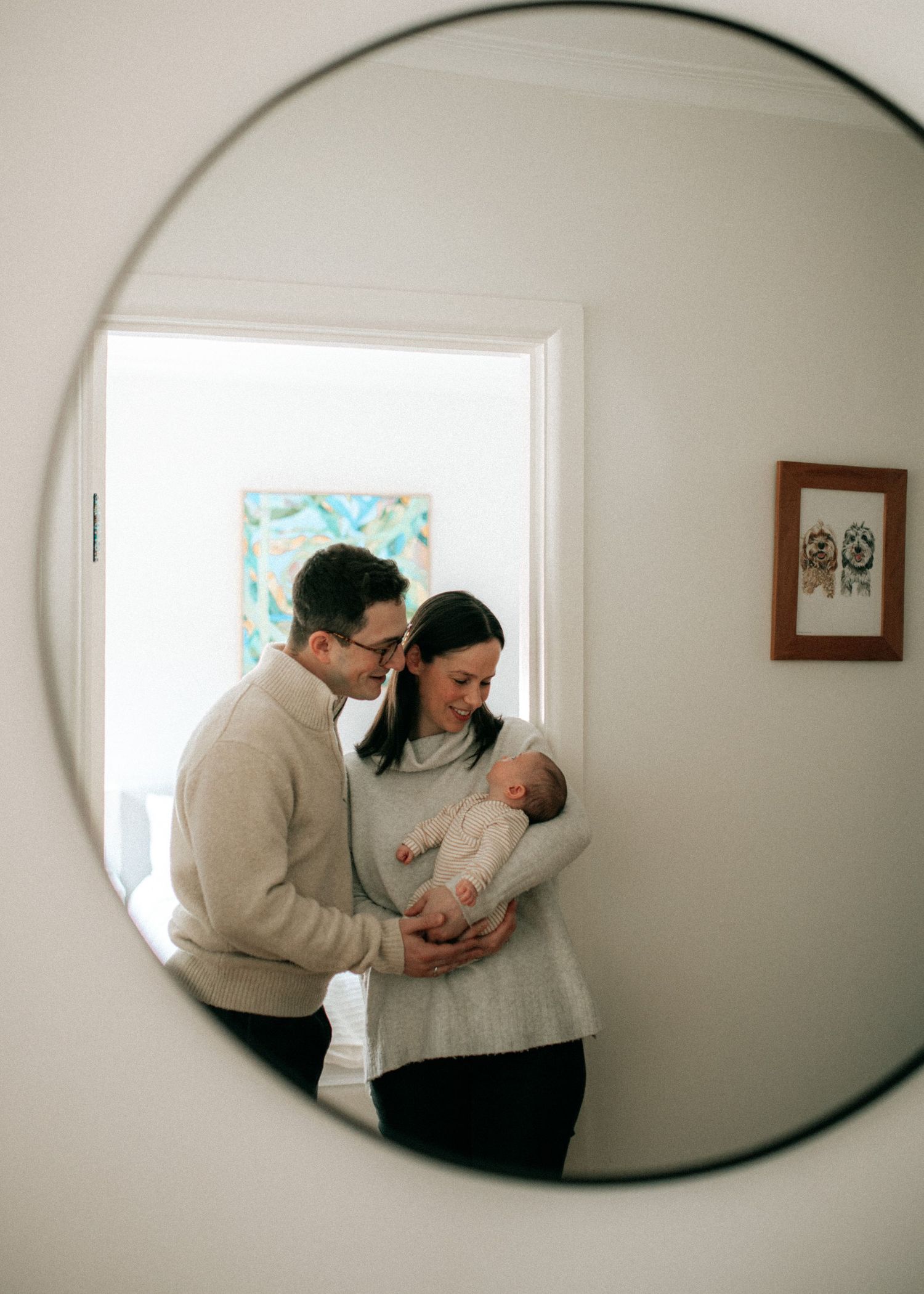 New parents admire their newborn baby in a round mirror reflection in a bright white home.