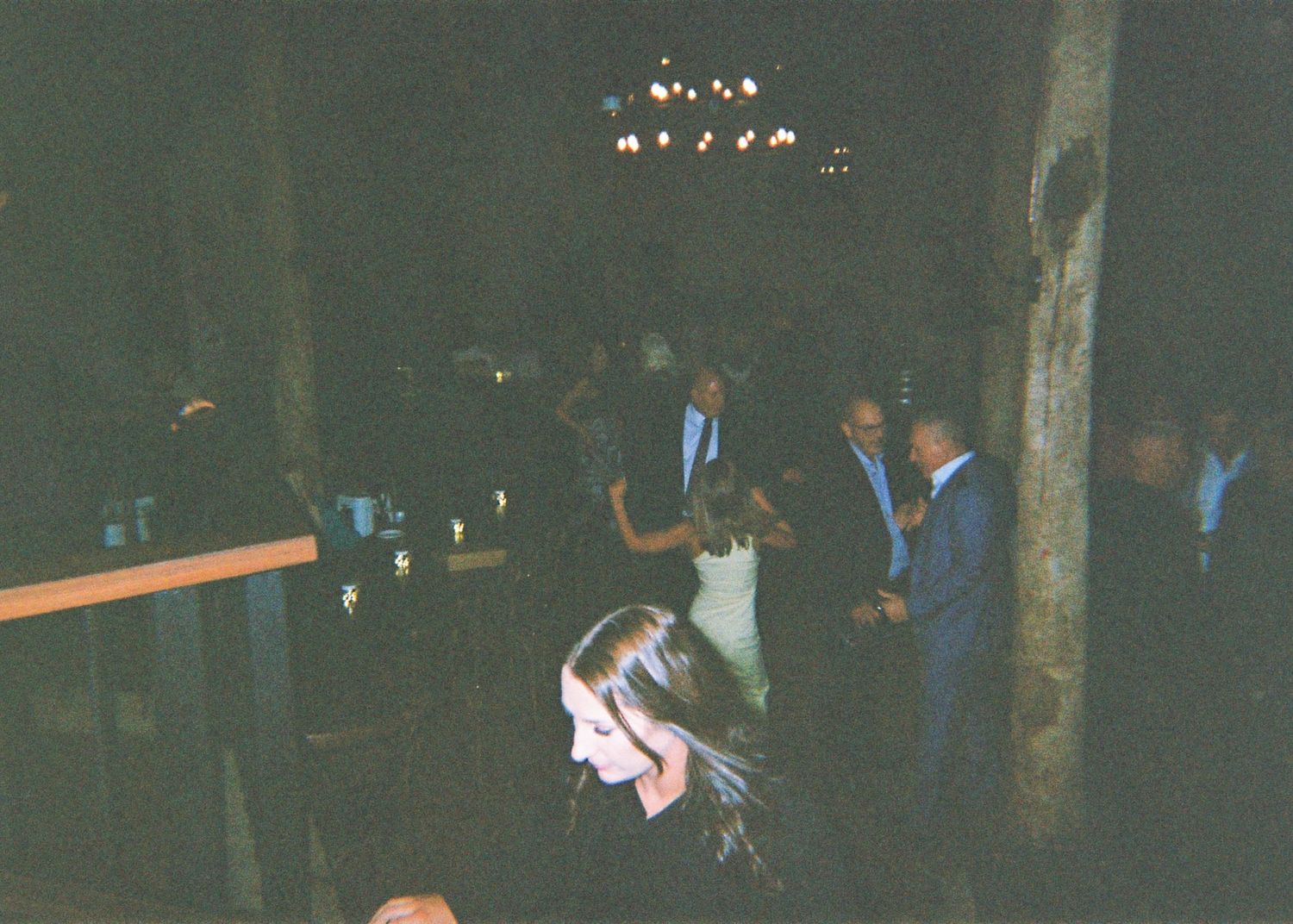 Wedding reception guests mingling and dancing in a dark rustic barn venue with warm lighting.