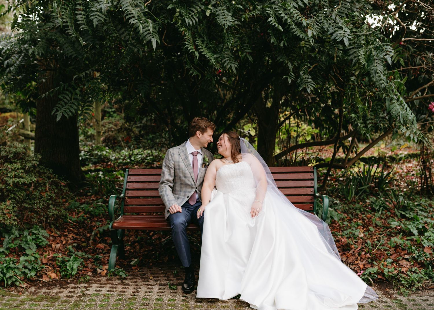 Bride and groom sitting on a wooden bench surrounded by lush green garden foliage.