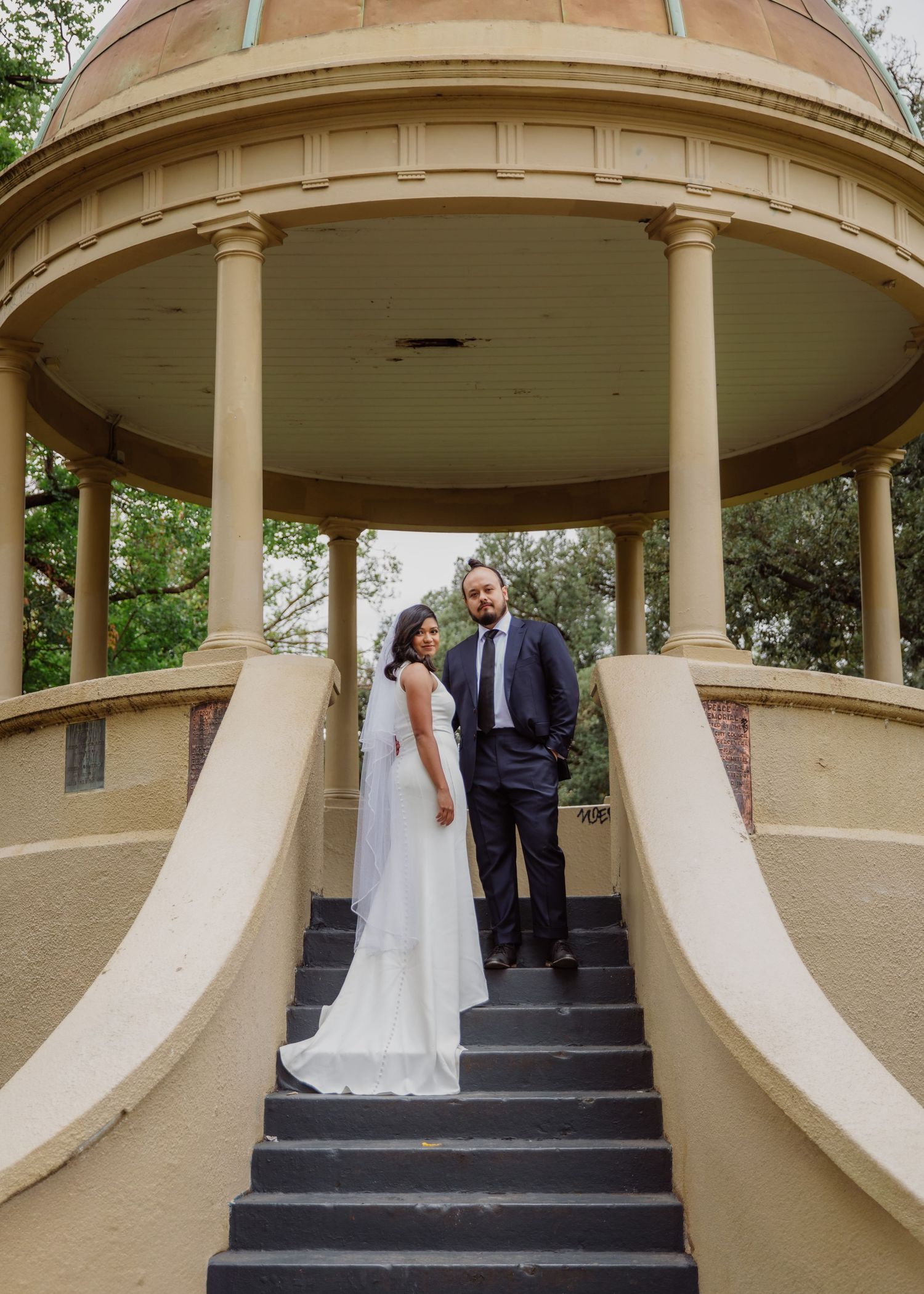 Bride and groom pose elegantly on steps of a classical rotunda gazebo in a park.