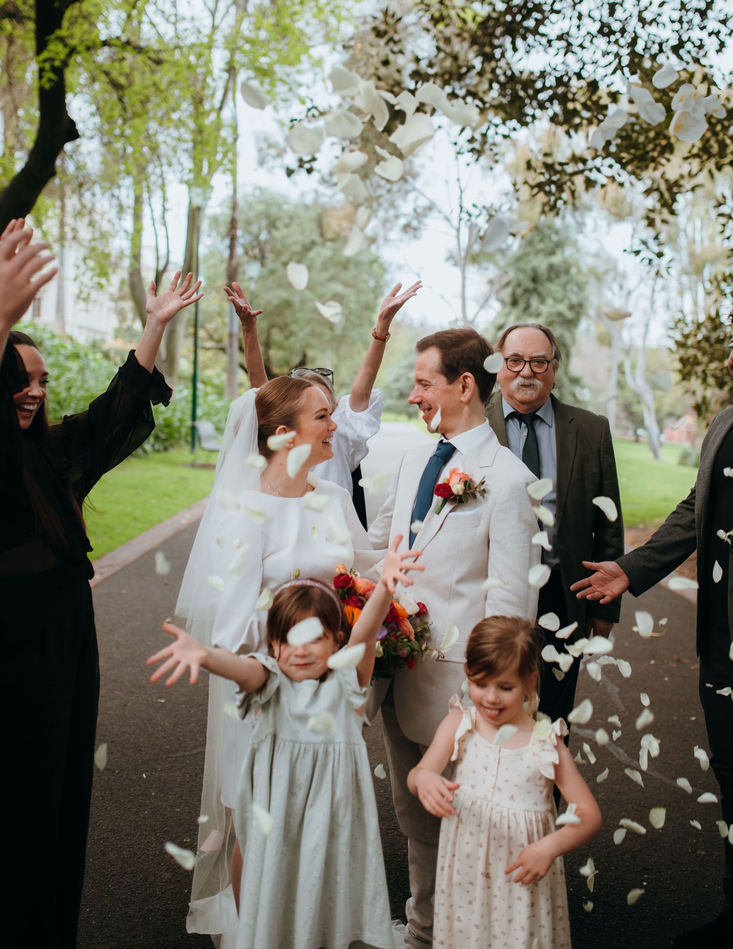 Wedding guests toss white flower petals over joyful bride and groom in a sunlit park.