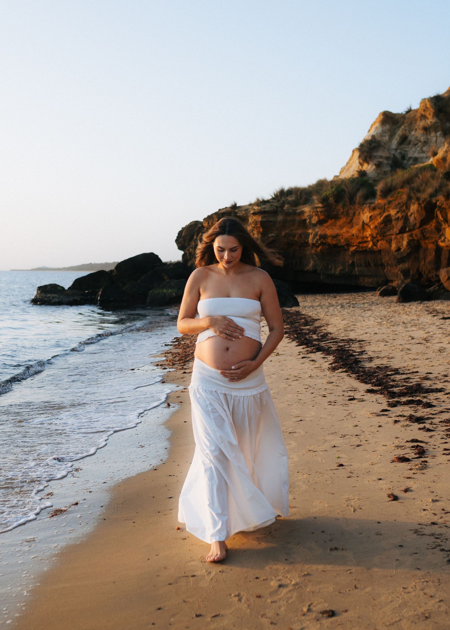 Pregnant woman in white outfit walking barefoot on a beach at golden hour near rocky cliffs.