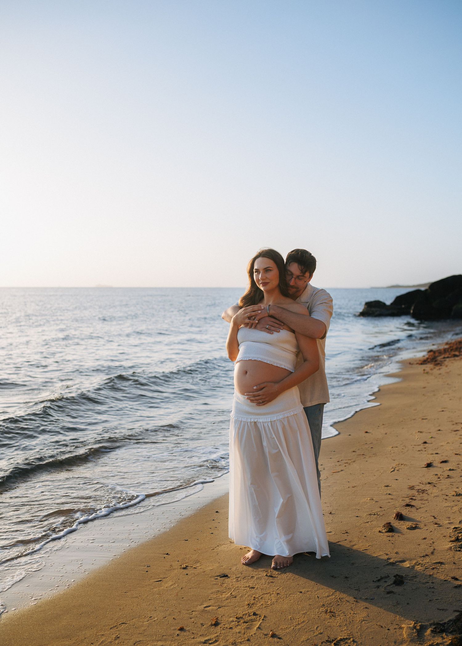 Couple sharing tender embrace on sandy beach at sunset, woman in flowing white maternity dress.