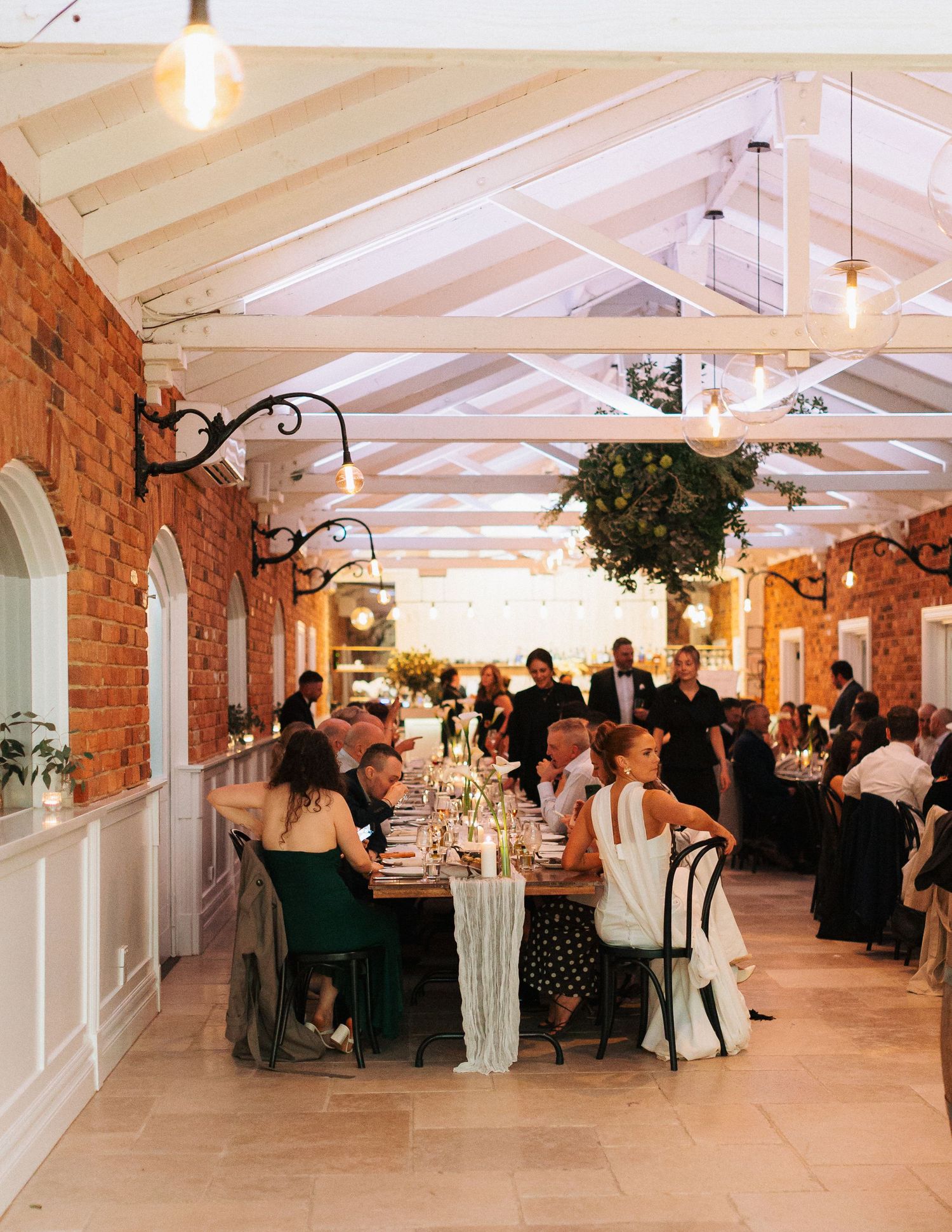 Elegant wedding reception in a brick conservatory venue with long dining tables, string lights, and guests seated for dinner.