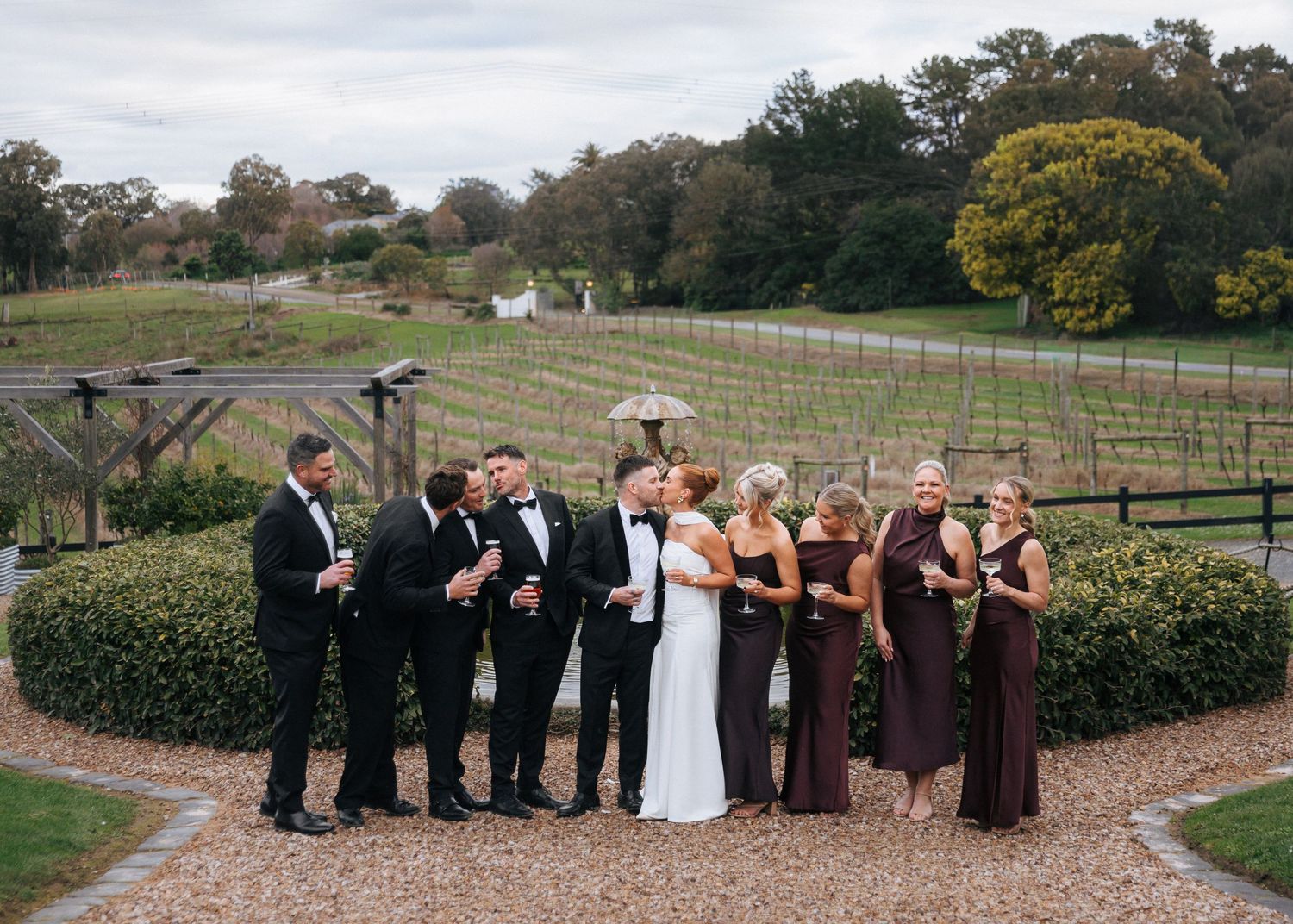 Wedding party in formal attire poses at a vineyard, with bridesmaids in burgundy gowns and groomsmen in black tuxedos.