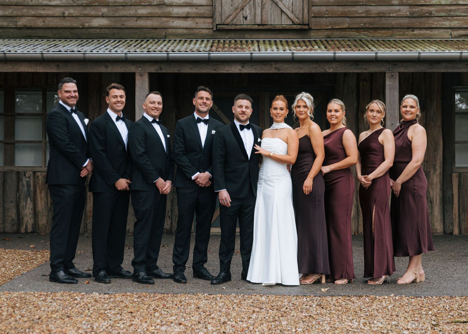 Wedding party posing together outdoors, bride in white gown with groomsmen in black tuxedos and bridesmaids in burgundy dresses.