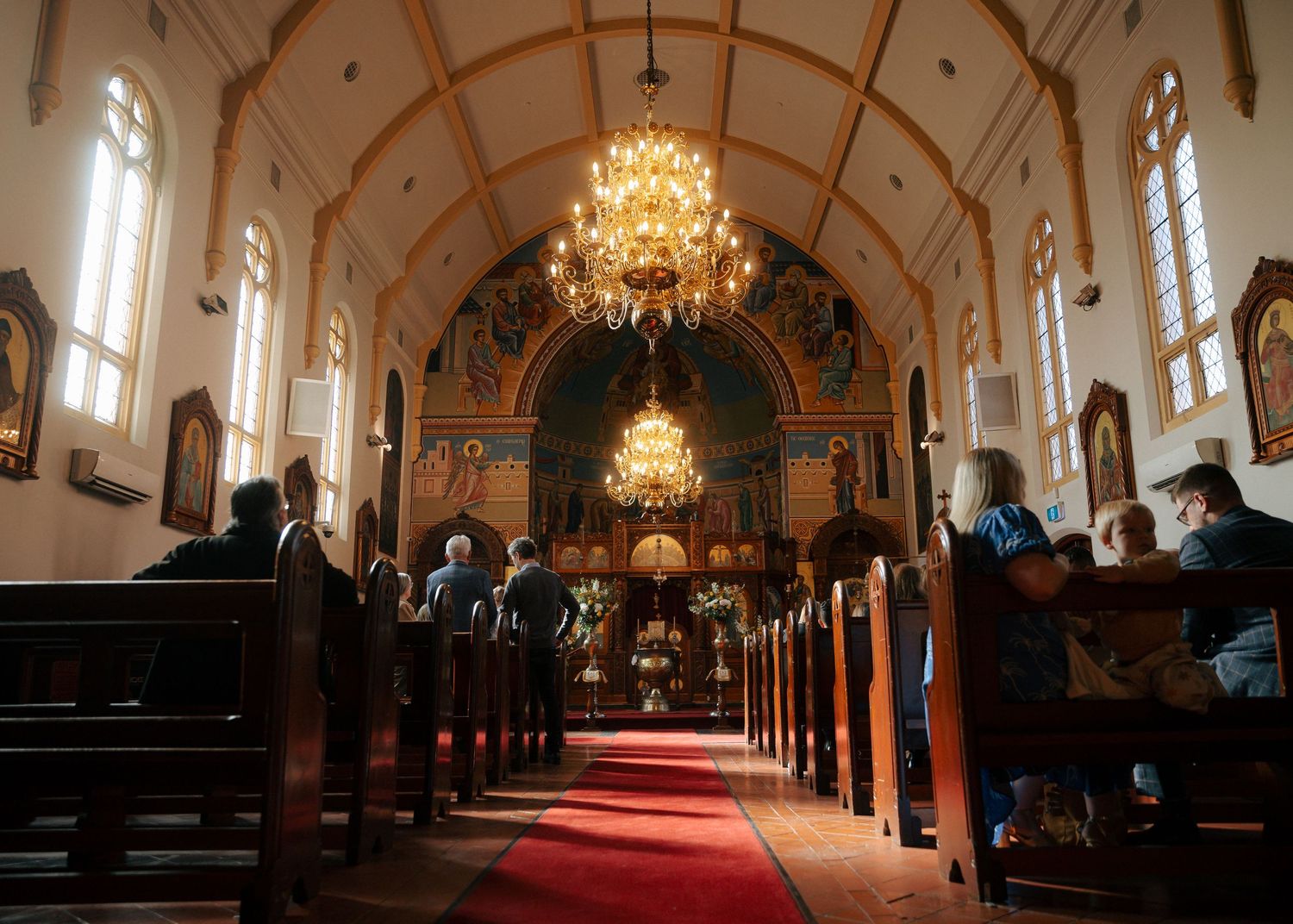 Interior of an ornate Orthodox church with chandeliers, red carpet aisle, and worshippers seated in wooden pews..jpg