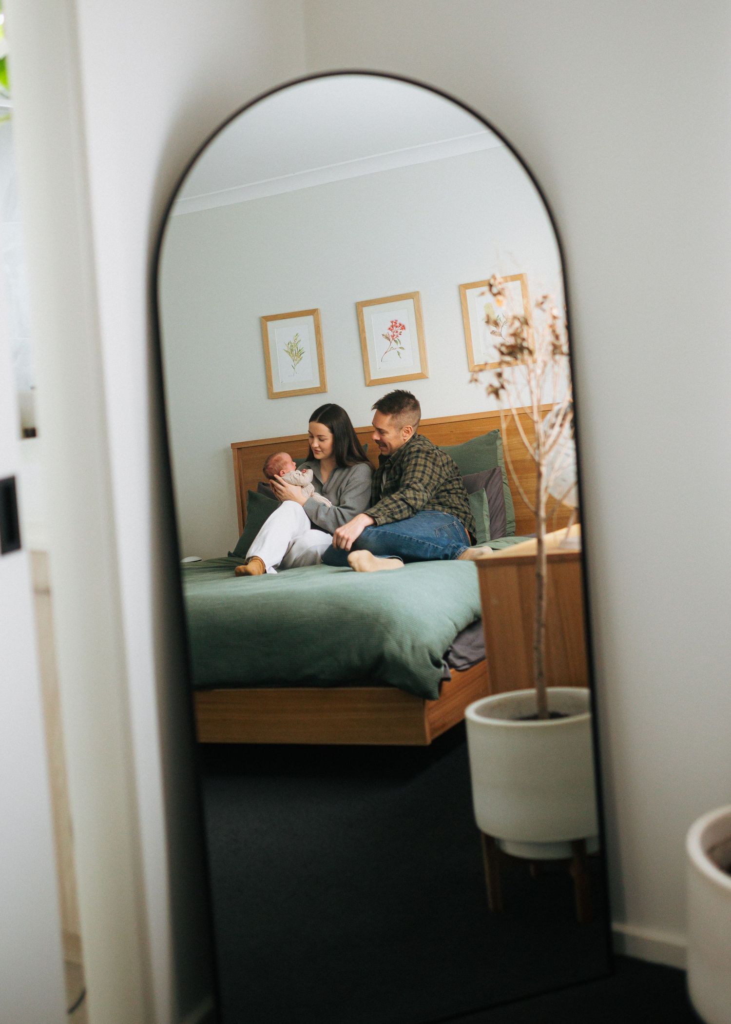 Mirror reflection of new parents sitting on a green bed smiling at their newborn baby.