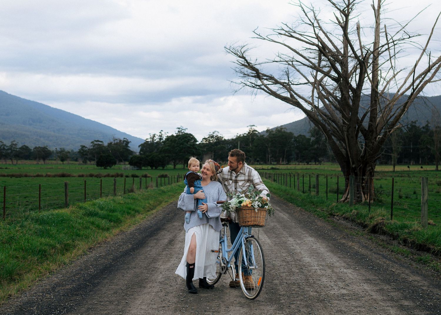 Happy family with toddler and vintage blue bicycle on a scenic rural gravel road.