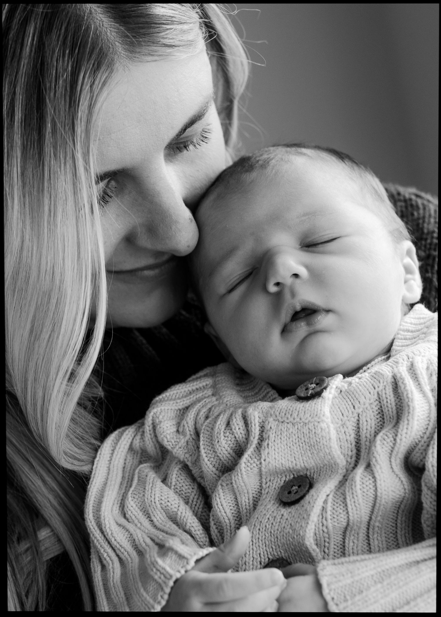 Black and white close-up of mother nuzzling sleeping newborn baby in cream knit cardigan.