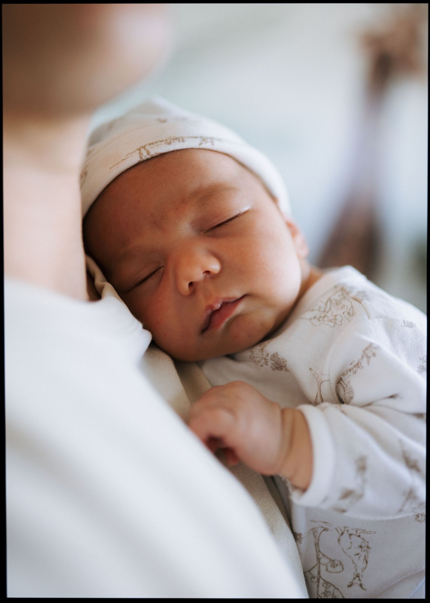 Sleeping newborn baby in a white cap and printed onesie rests peacefully against an adult's shoulder.