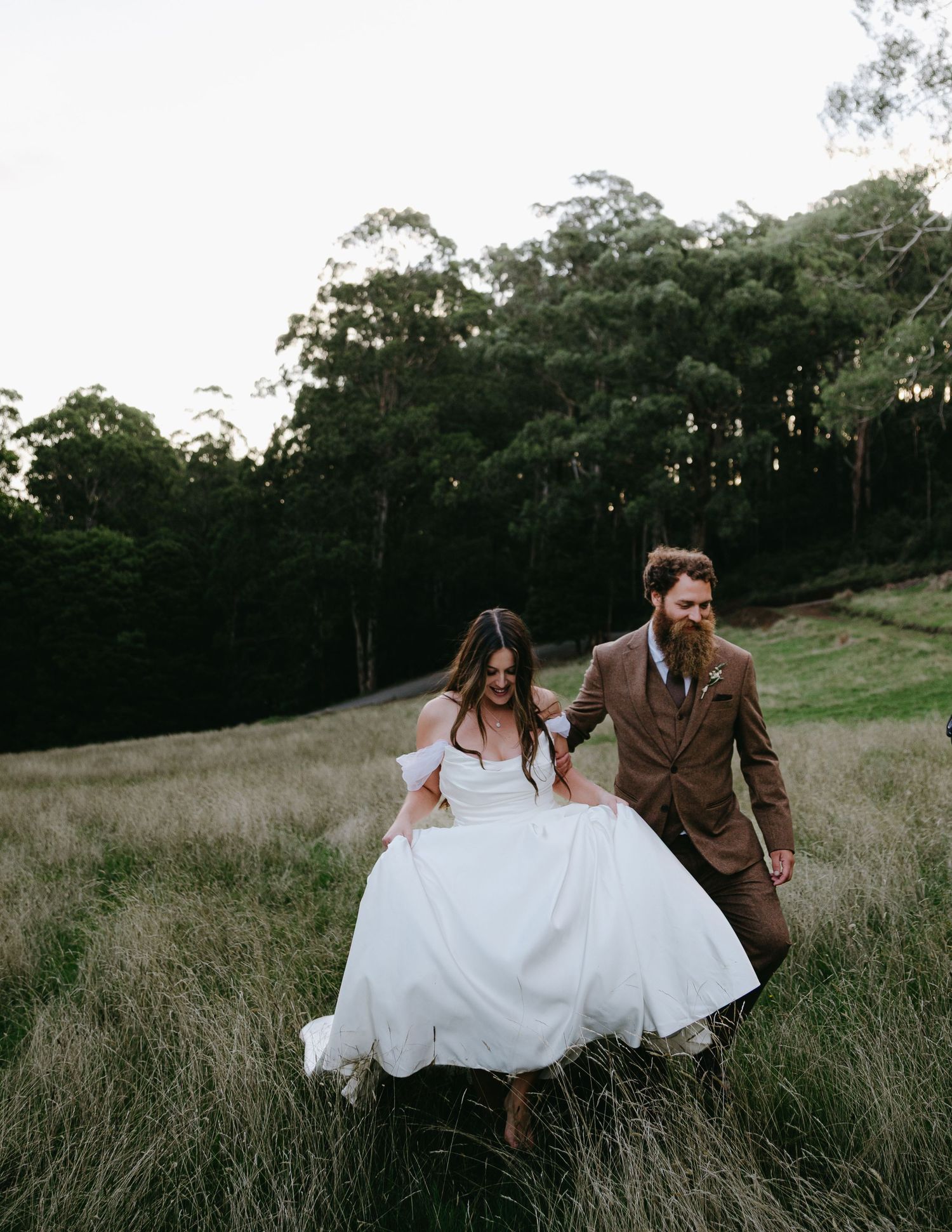 Bride and groom laughing as they walk through golden meadow grass, bride lifting white wedding gown.