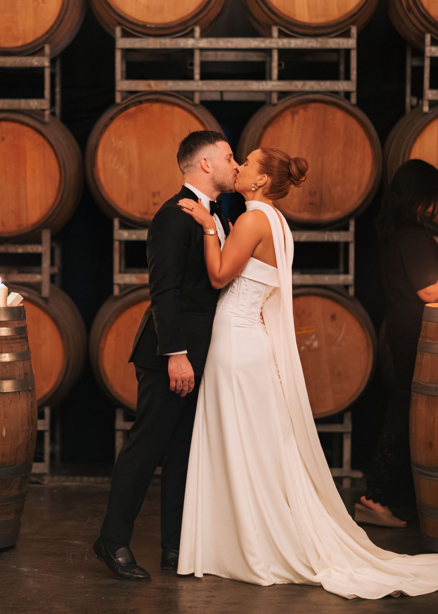 Bride and groom sharing first kiss surrounded by oak wine barrels in winery.