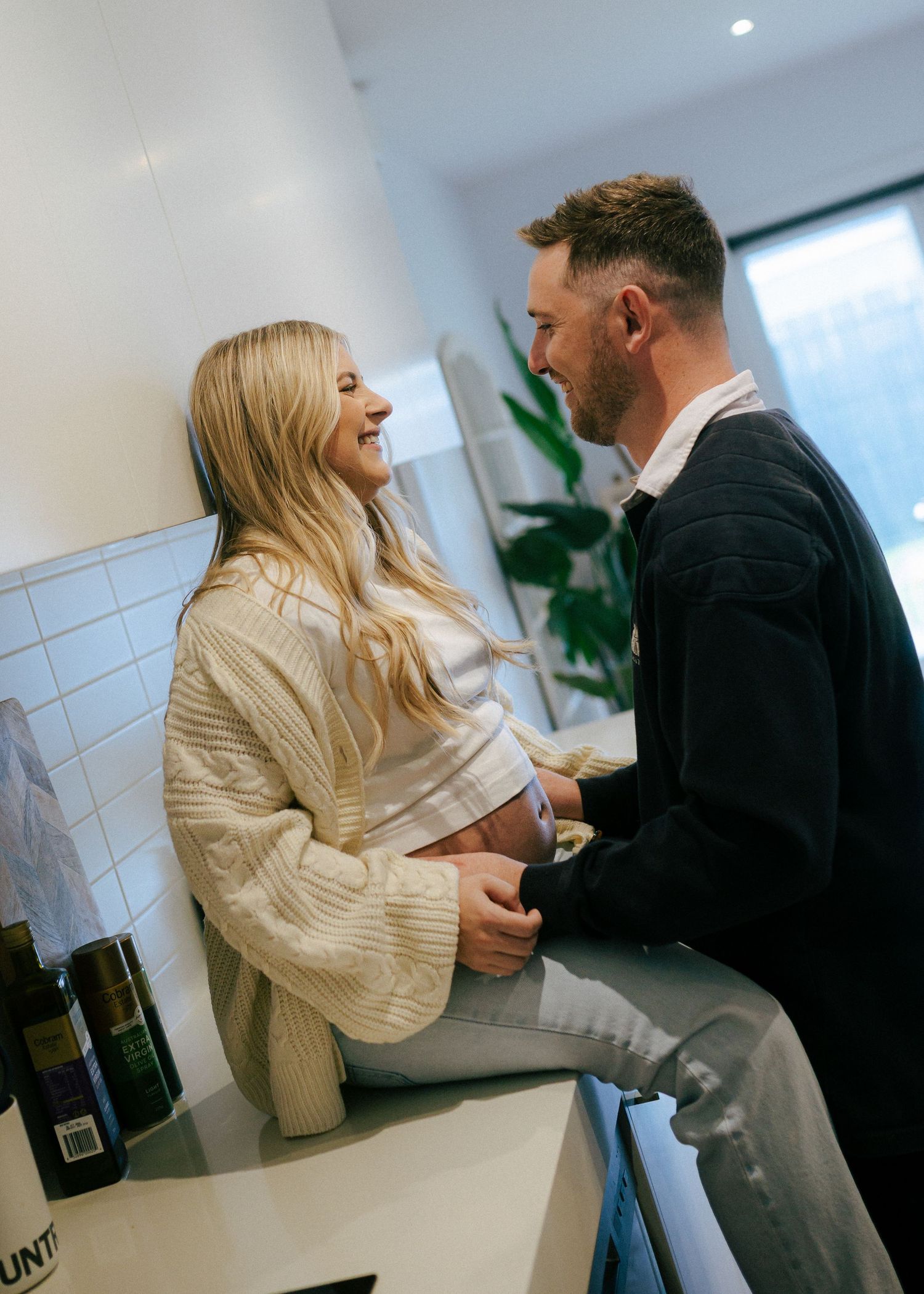 Smiling pregnant couple sharing tender moment in modern kitchen, man touching baby bump.
