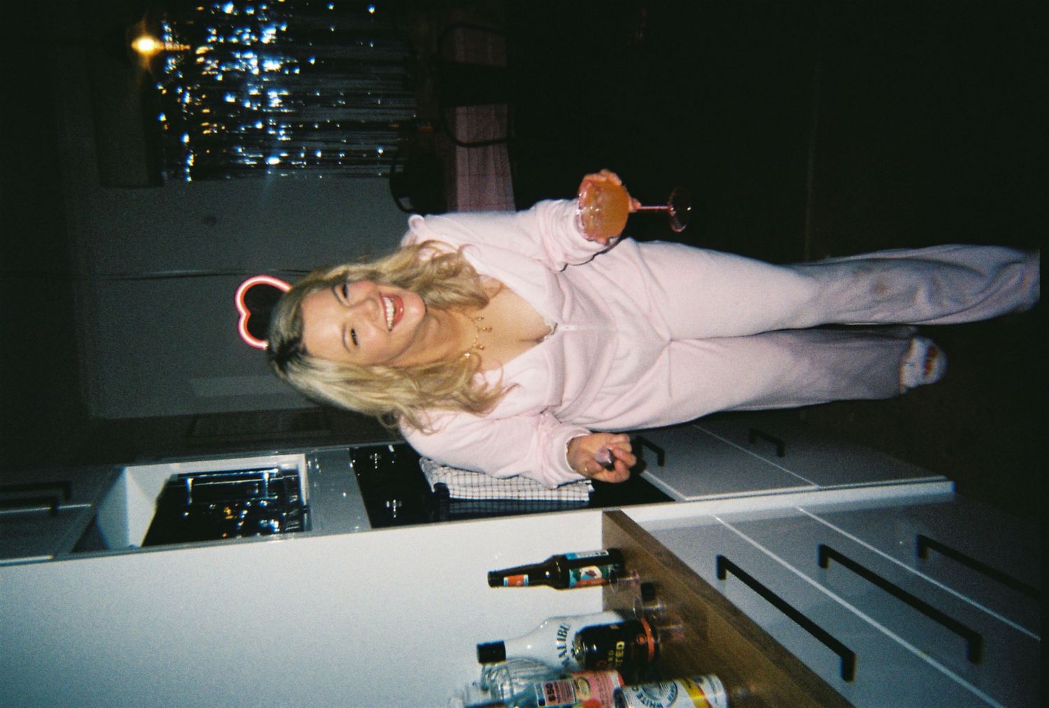 A smiling blonde woman in a pink outfit wearing devil horns poses at a kitchen counter at a party.