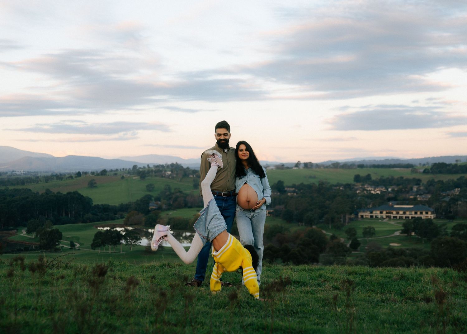 Joyful family maternity photo with child doing cartwheel on scenic green hillside.