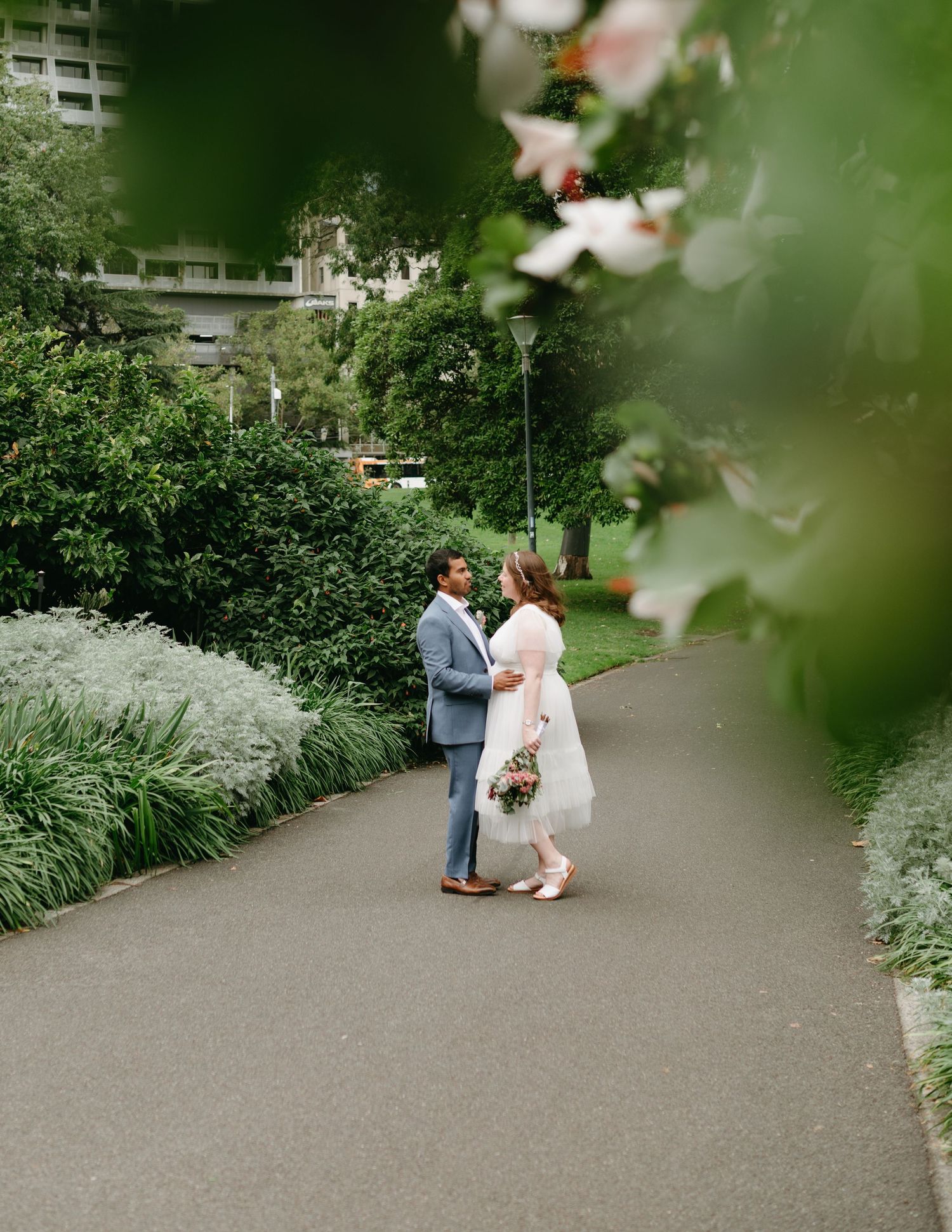 Wedding couple stands on a garden path surrounded by lush greenery and blooming flowers during their outdoor portrait session.