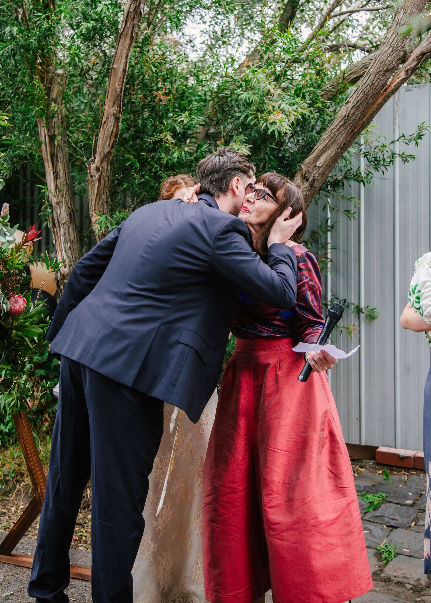 Groom in navy suit embraces woman in red dress near rustic wooden arch at garden wedding.