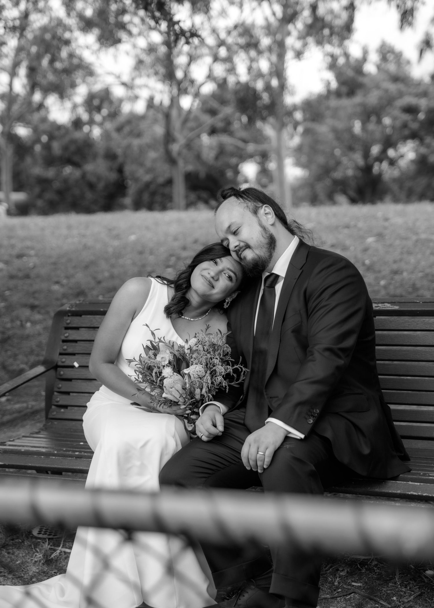 Black and white photo of bride and groom sitting intimately on a park bench.