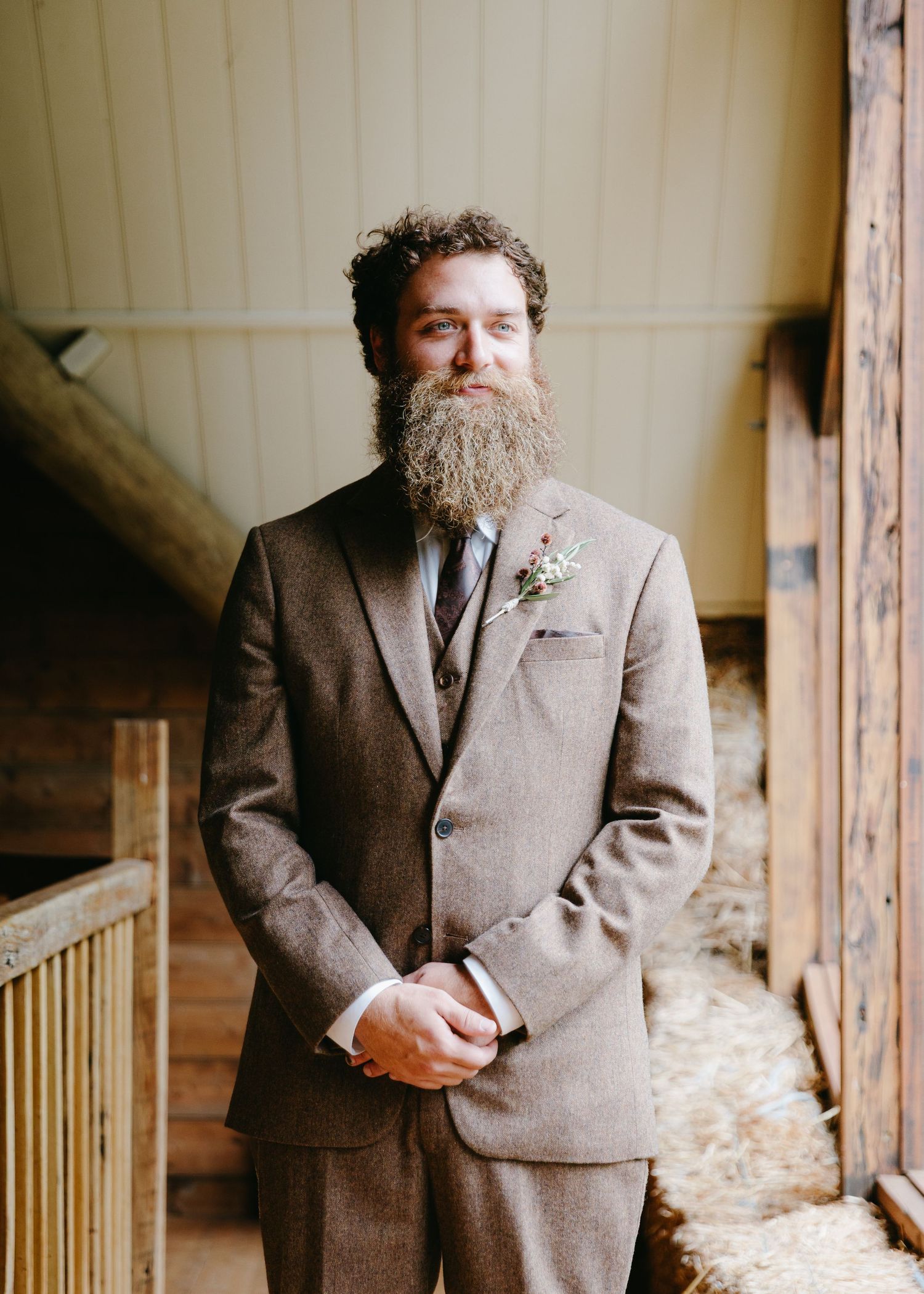 Bearded groom in a brown tweed three-piece suit with a floral boutonniere standing in a rustic barn.
