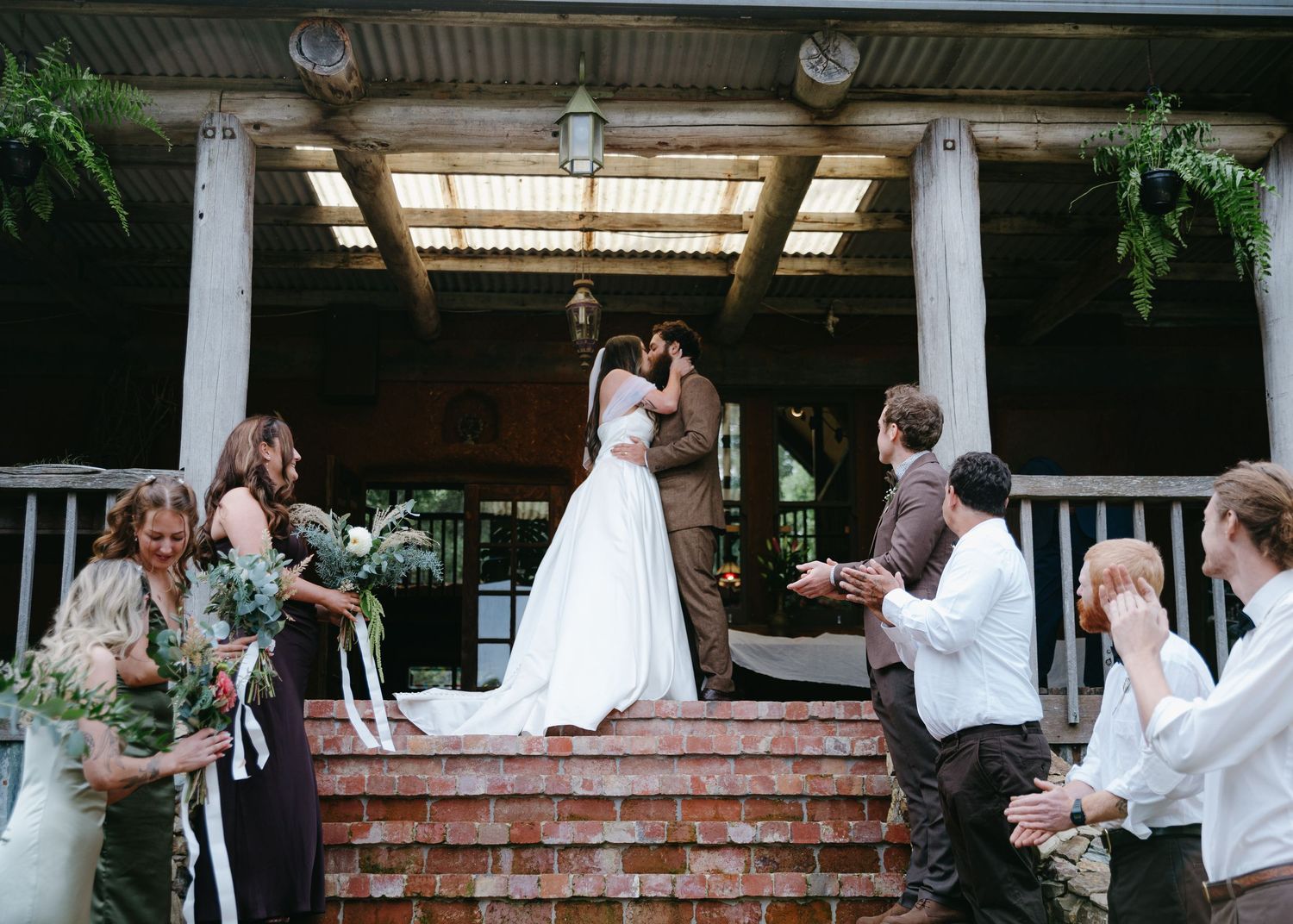 Bride and groom kissing on brick steps surrounded by cheering wedding party at outdoor venue.