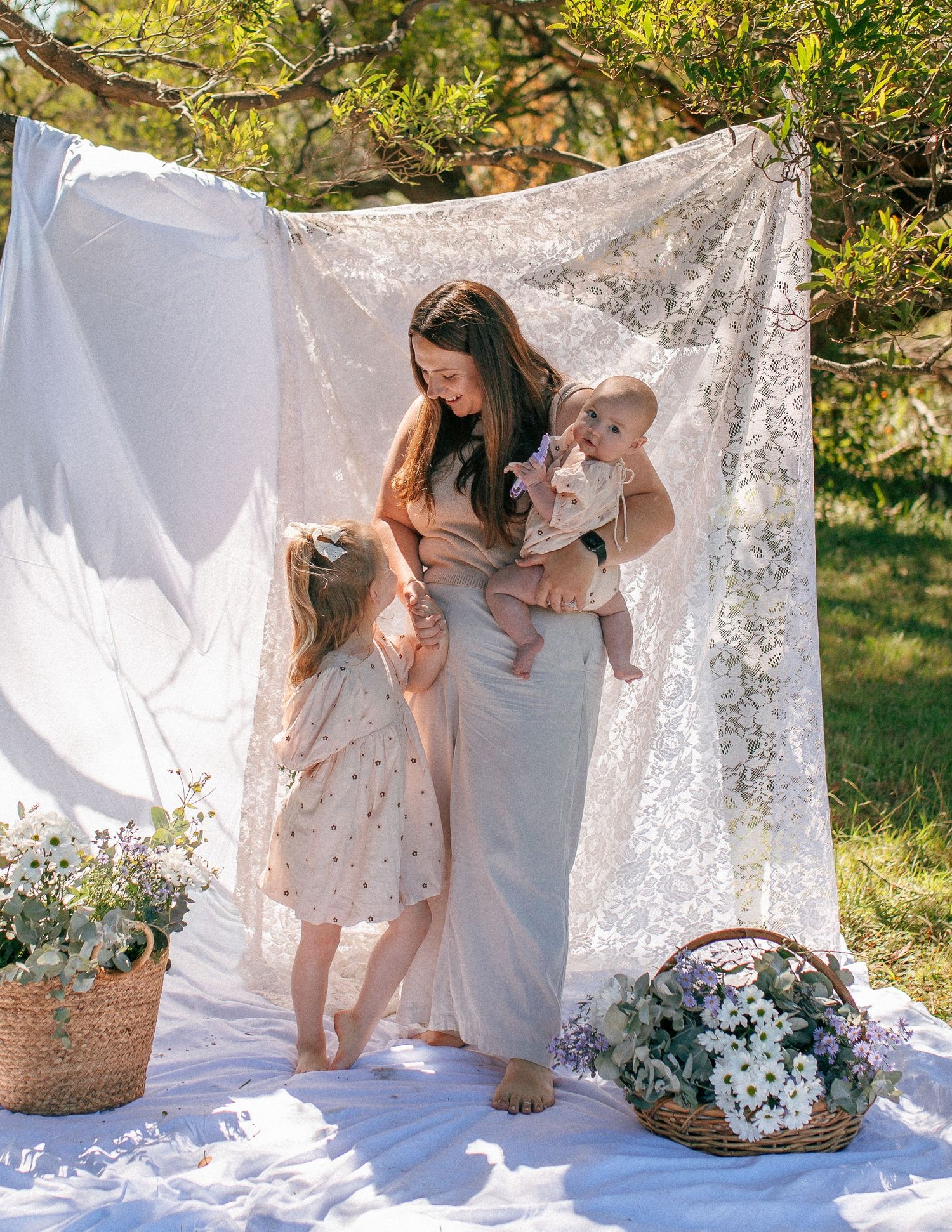 A mother holds her baby while her toddler daughter stands beside her in front of a lace backdrop outdoors.