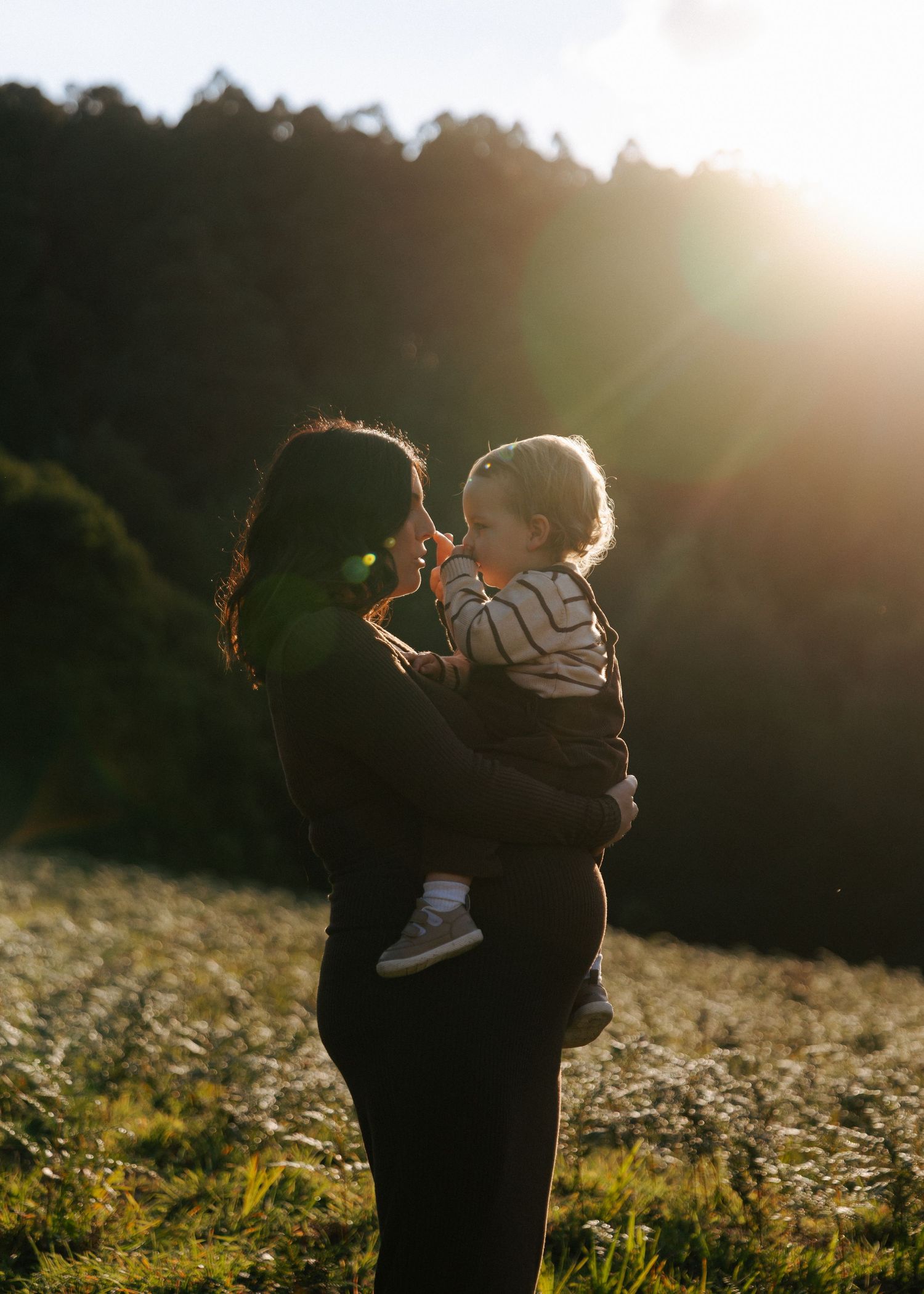 Mother holding toddler face-to-face in glowing golden sunset light in open meadow.